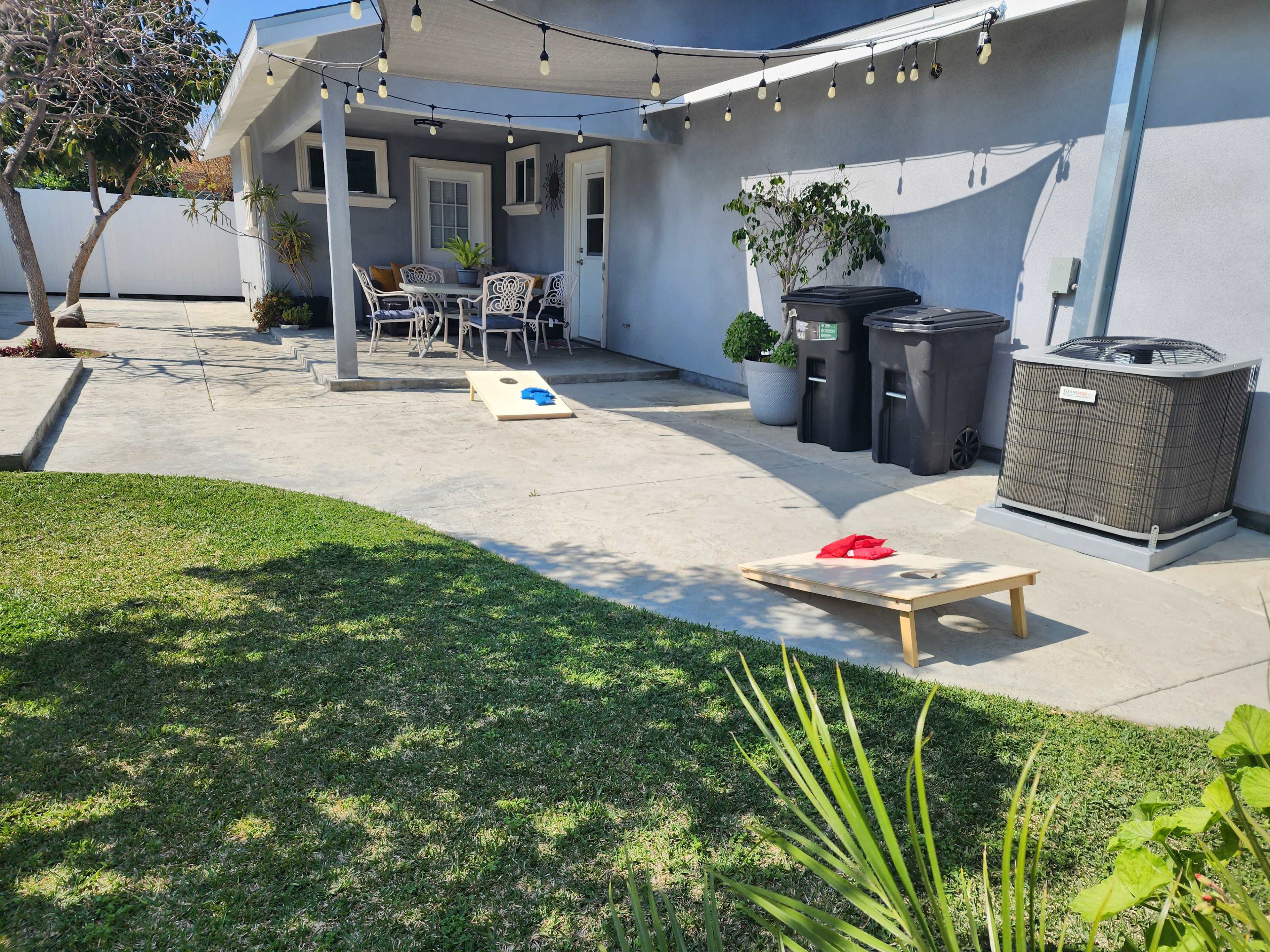 The image shows a backyard patio area with two cornhole boards, outdoor seating, trash bins, and an air conditioning unit.
