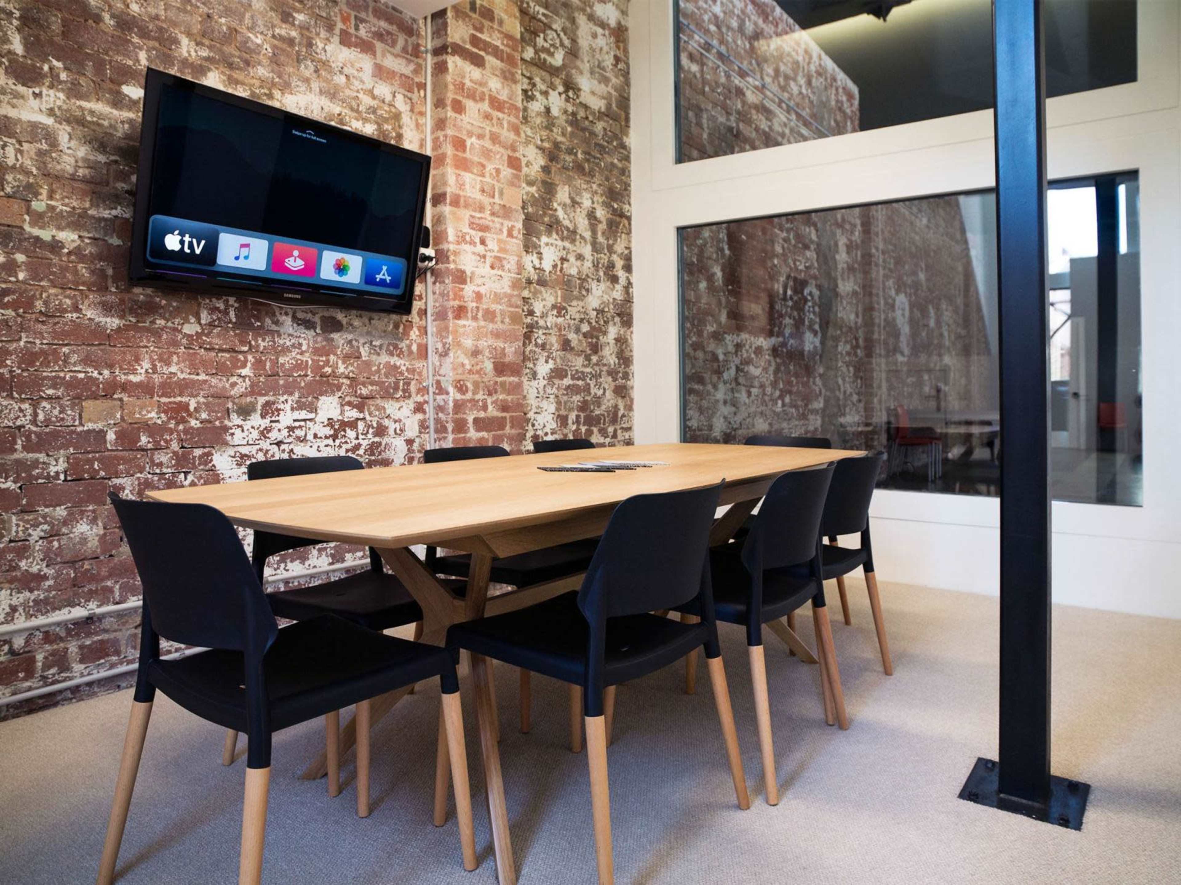 The image shows a modern meeting room with a wooden table surrounded by black chairs, a television mounted on a brick wall, and large windows letting in natural light.