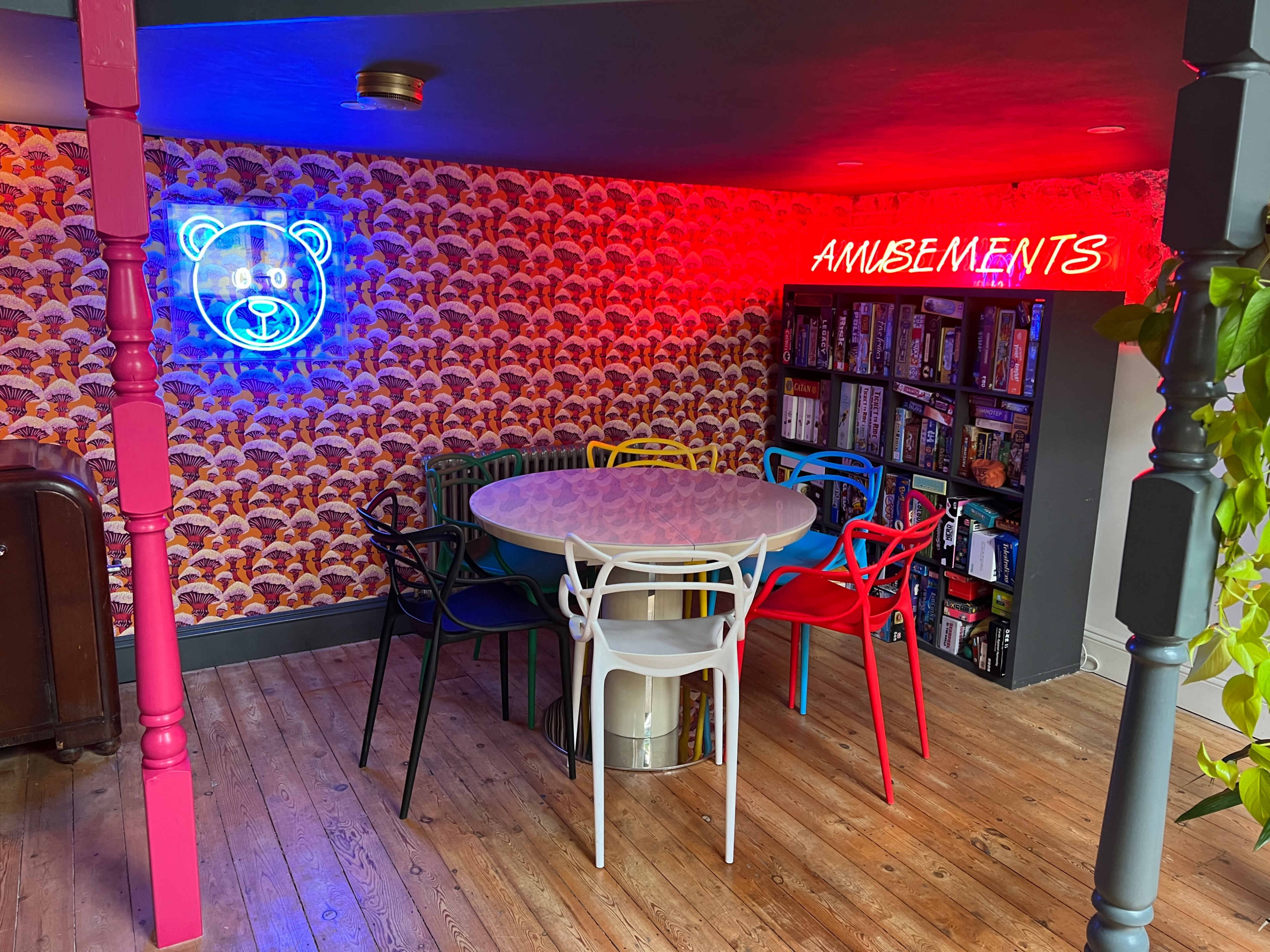 A colorful seating area features a round table surrounded by variously colored chairs against a patterned wallpaper backdrop, with a neon sign reading "AMUSEMENTS" illuminating the space.