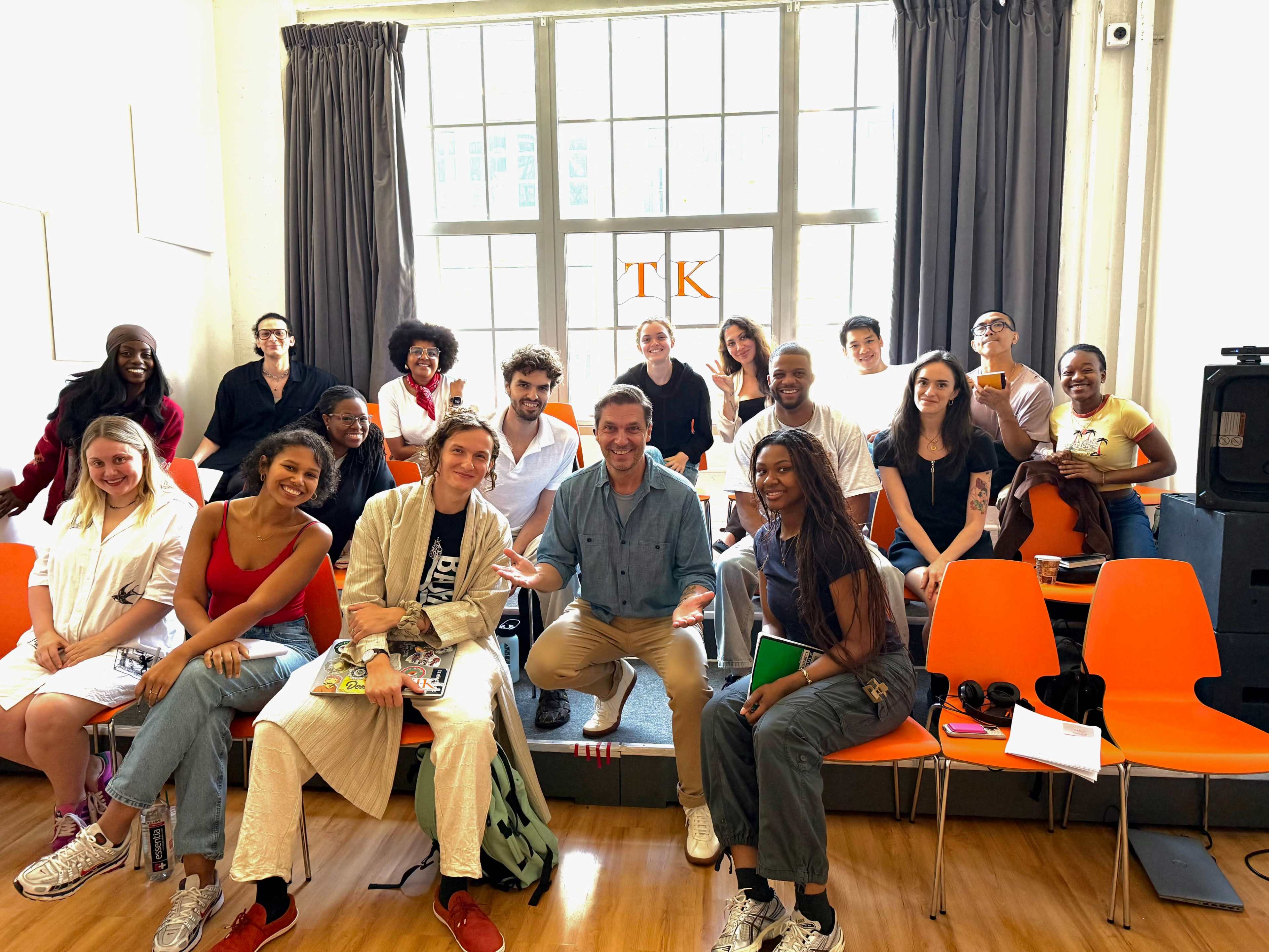 A diverse group of individuals is seated and standing in a brightly lit room with large windows, all facing the camera and smiling.