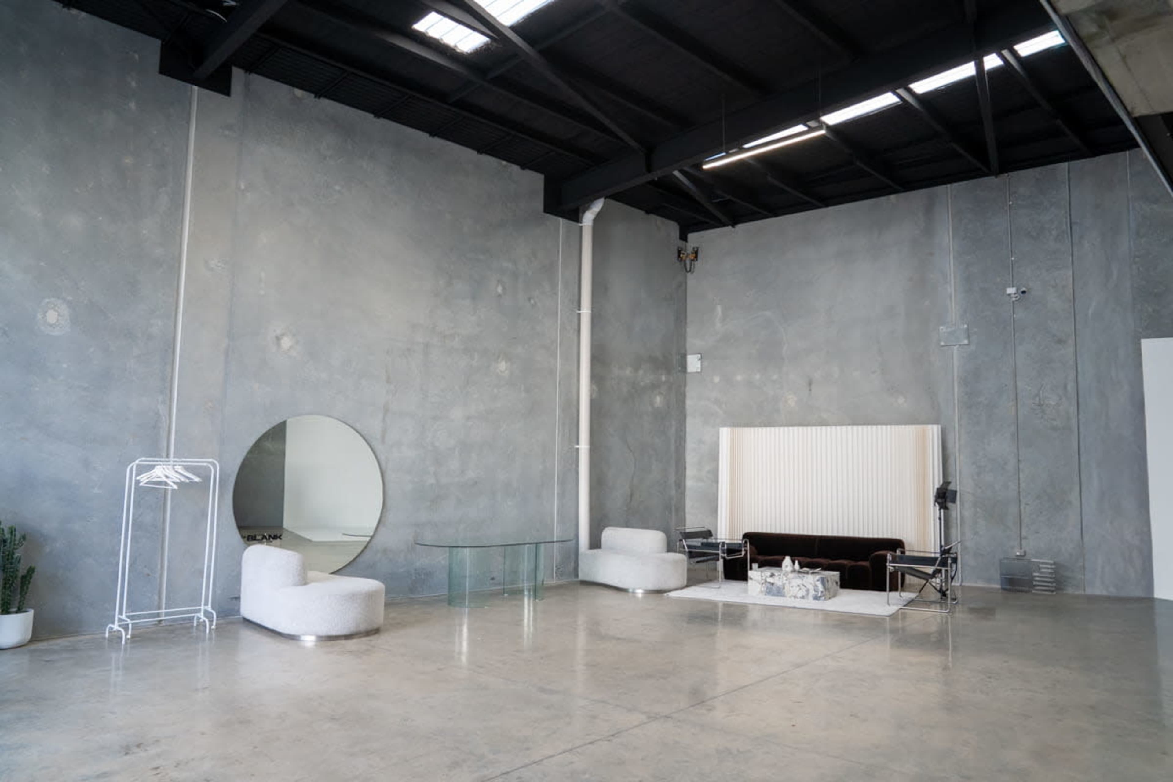 The image shows a minimalist indoor space with concrete walls, a glass table, a round mirror, a dark sofa, and light-colored seating arrangements.