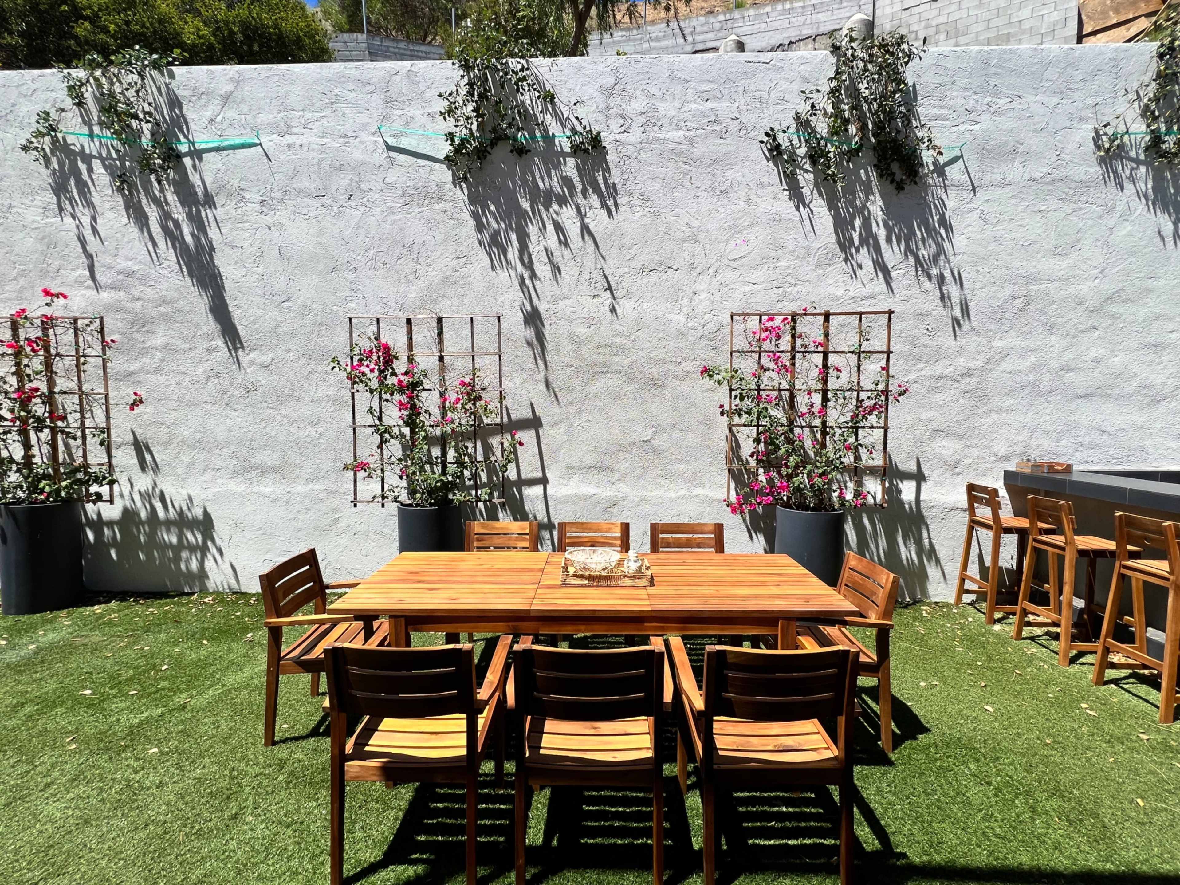A wooden dining table surrounded by chairs is set on a grassy area in front of a gray wall adorned with climbing plants and flower trellises.