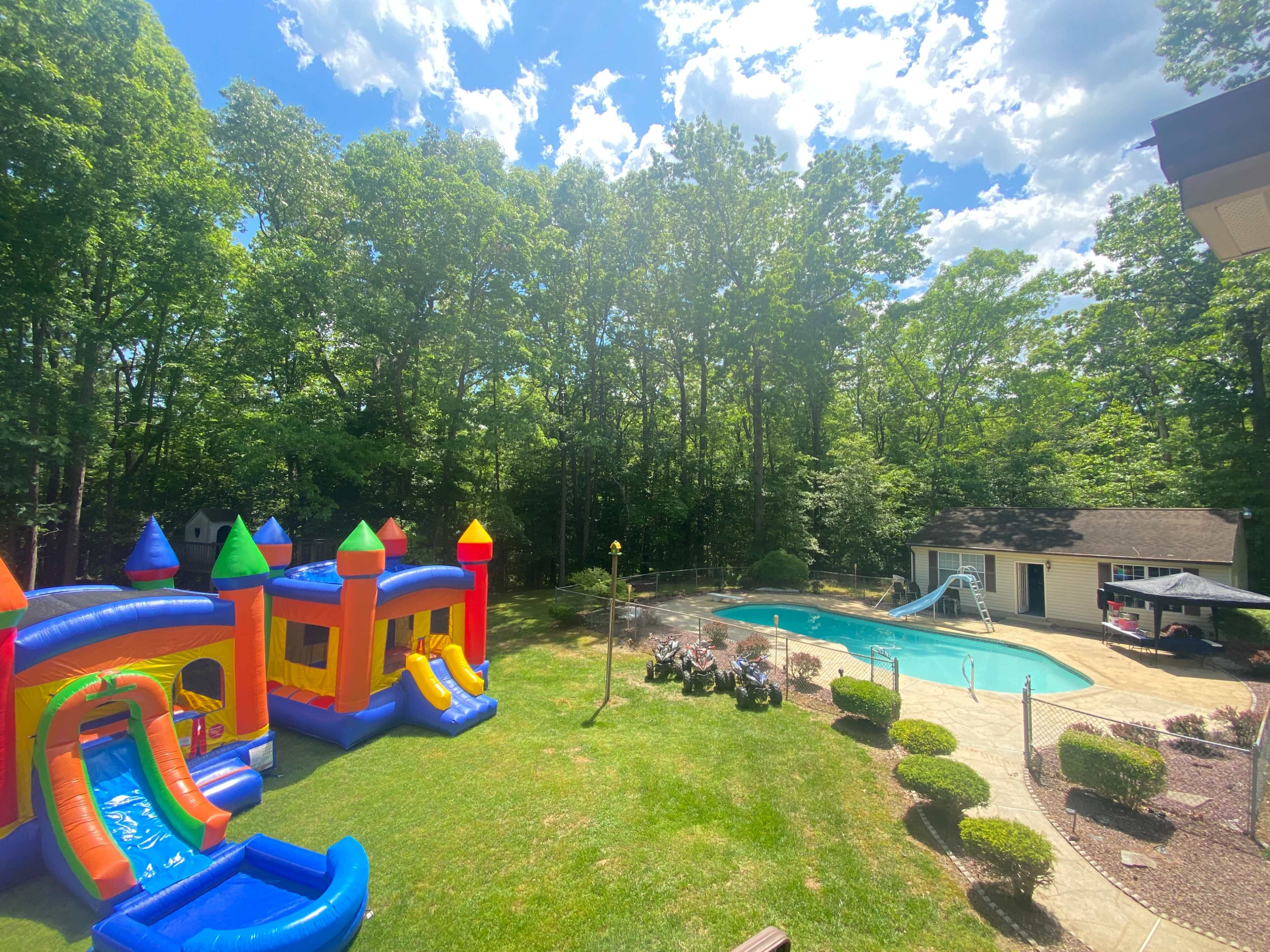 A backyard featuring a colorful inflatable bounce house and slide next to a swimming pool surrounded by trees.