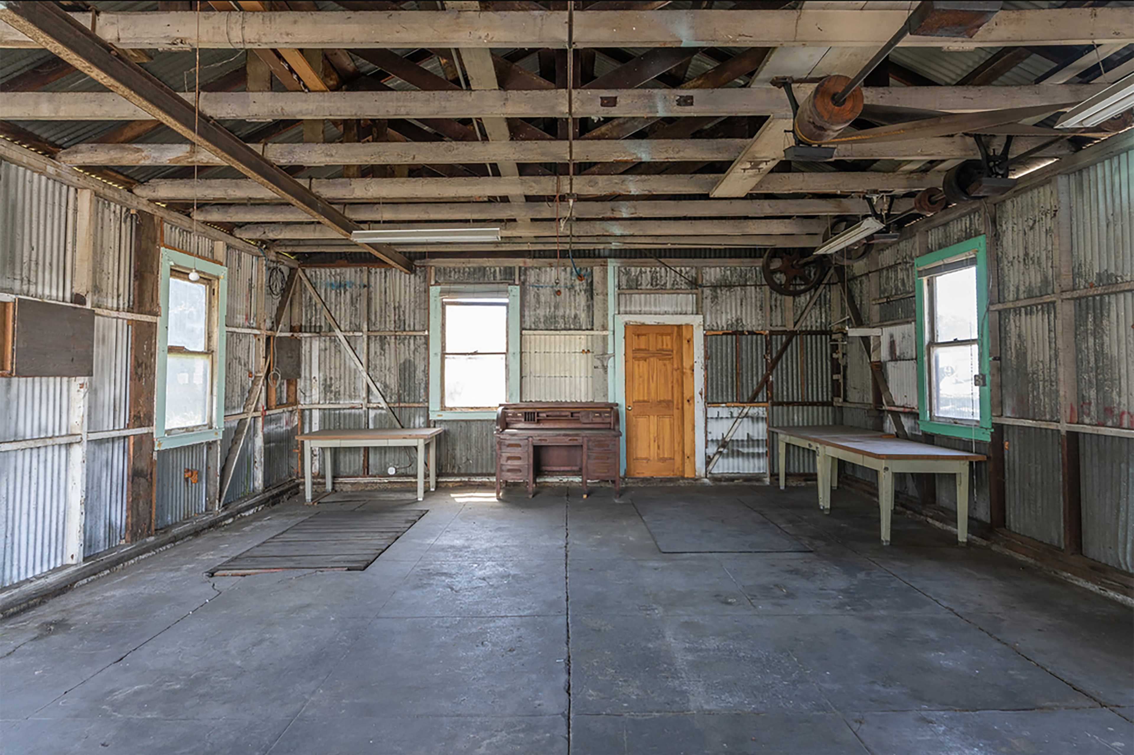 An empty, corrugated metal building interior with exposed beams, wooden tables, and two windows.