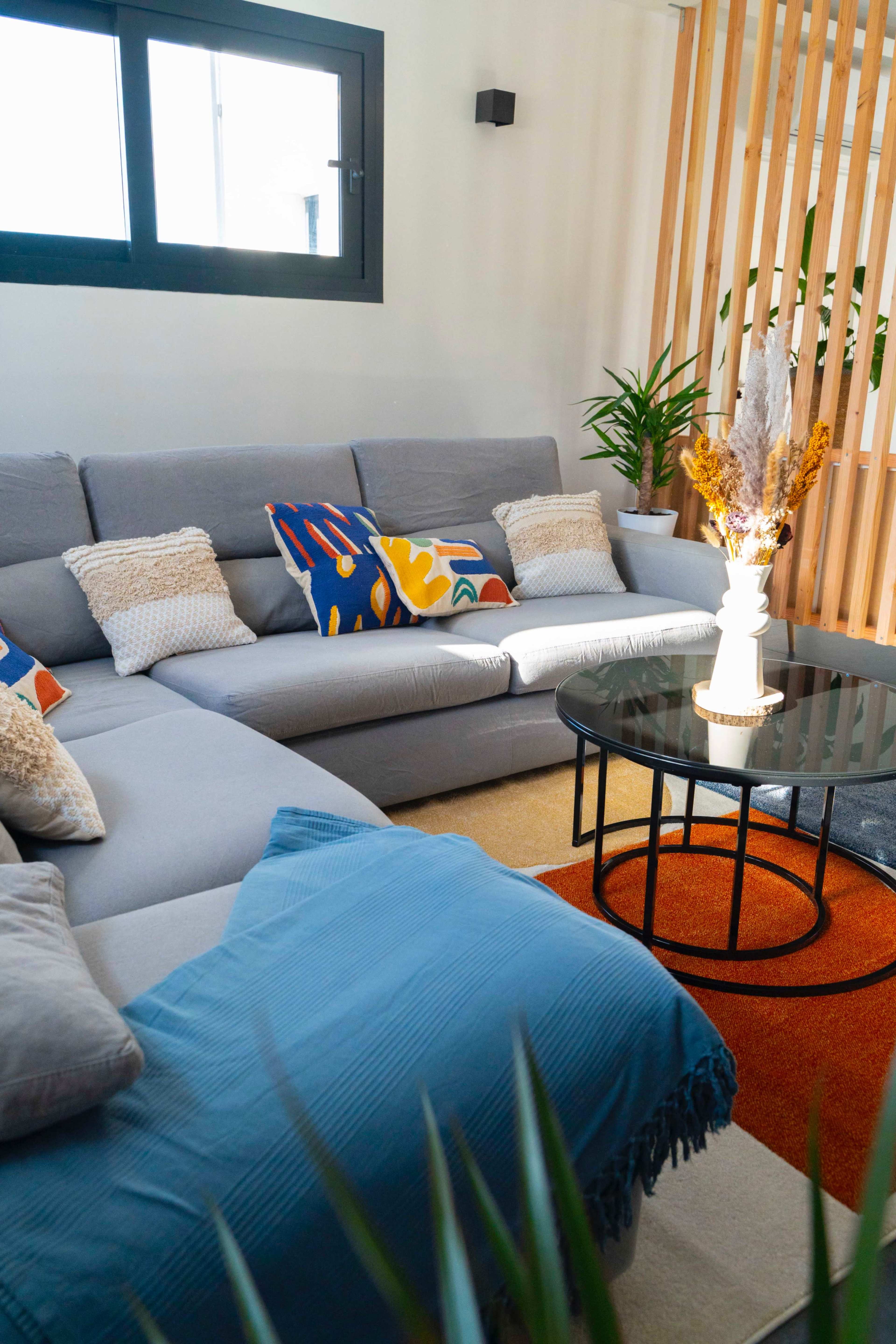 A modern living room features a gray sectional sofa with colorful throw pillows, a circular glass coffee table, and indoor plants beside a wooden slatted partition.