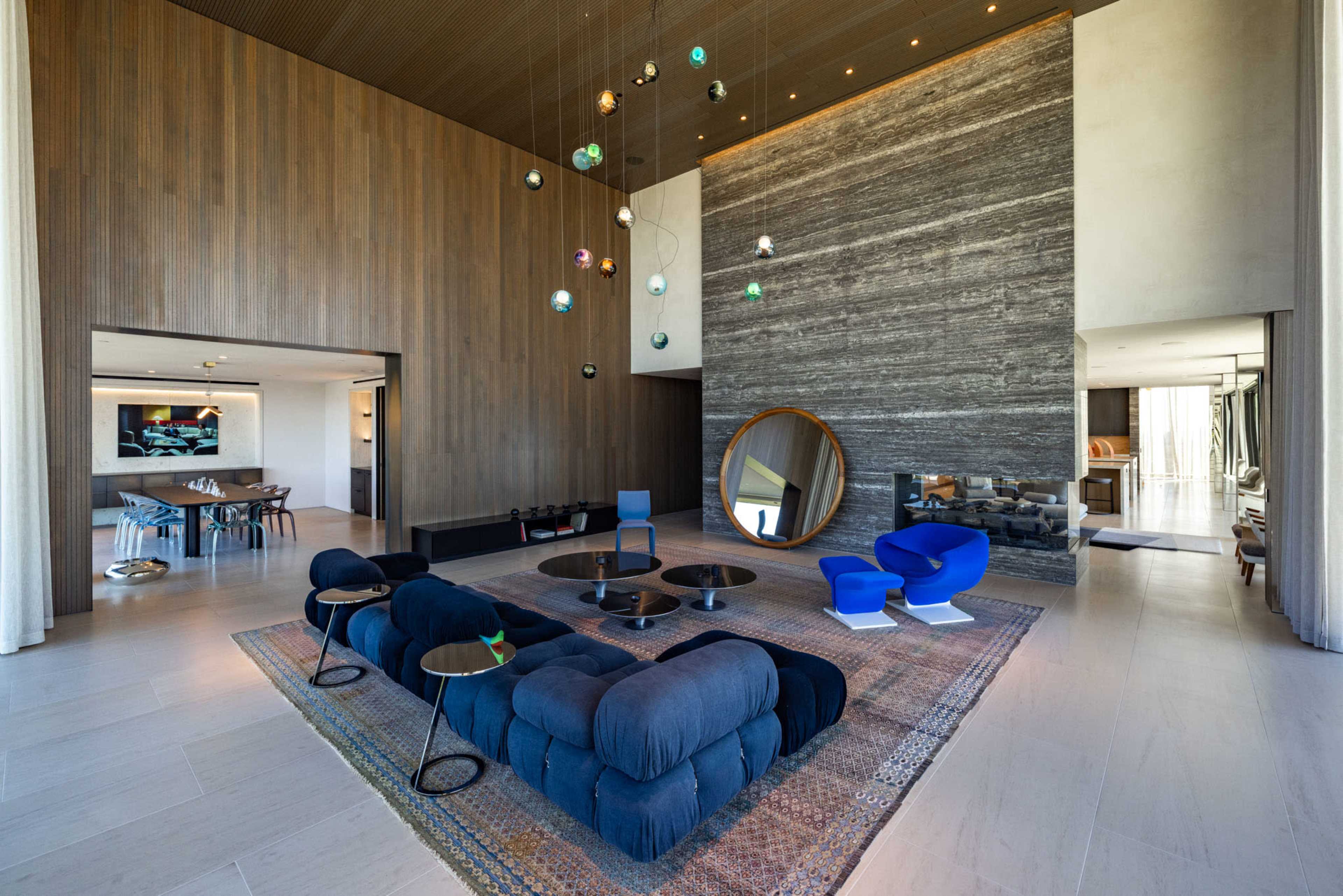 The image shows a modern living room with wooden walls, a stone feature wall, stylish furniture, and a large circular mirror.