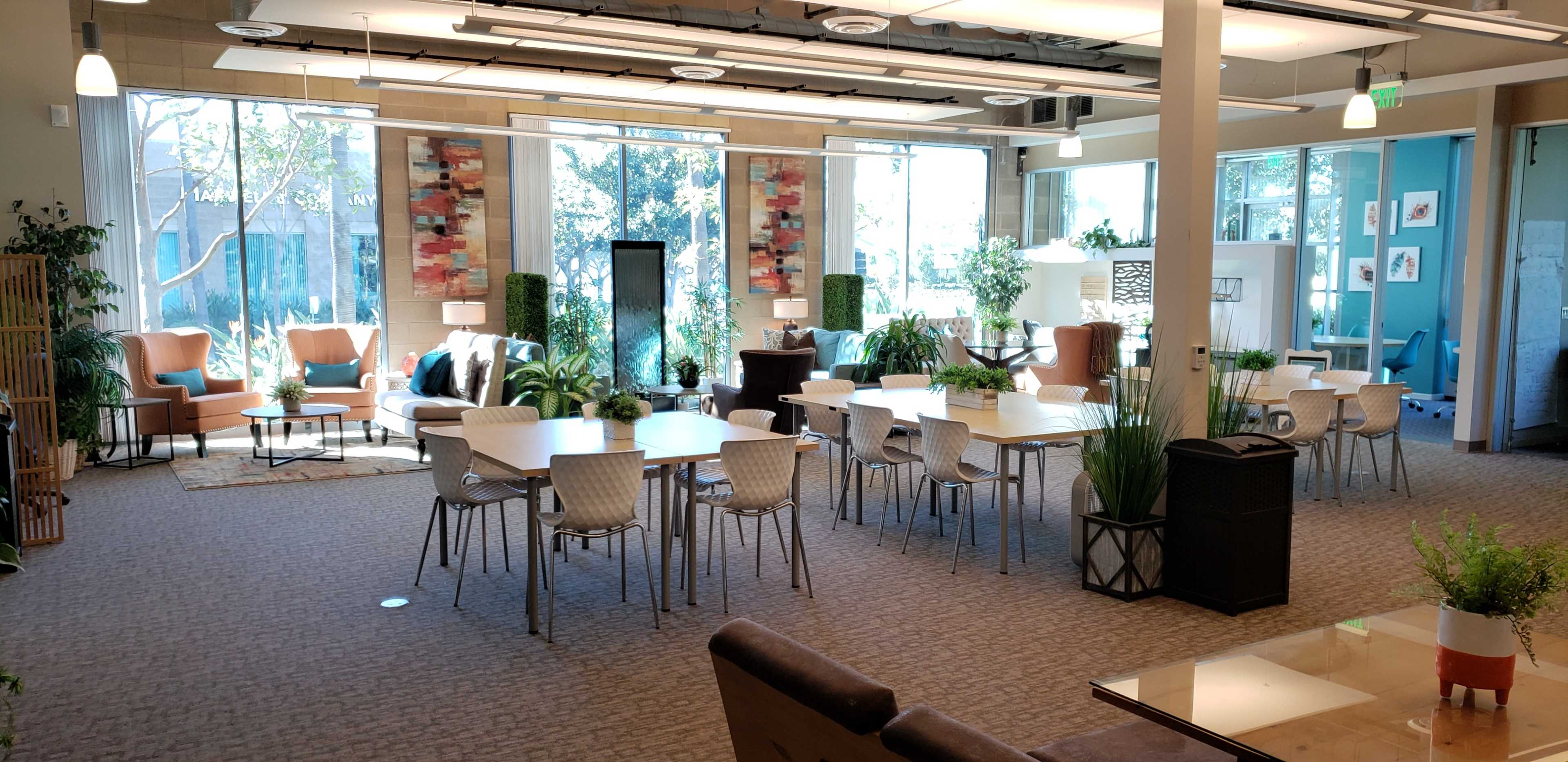 The image shows a spacious, well-lit common area with multiple seating arrangements, tables, and abundant greenery.