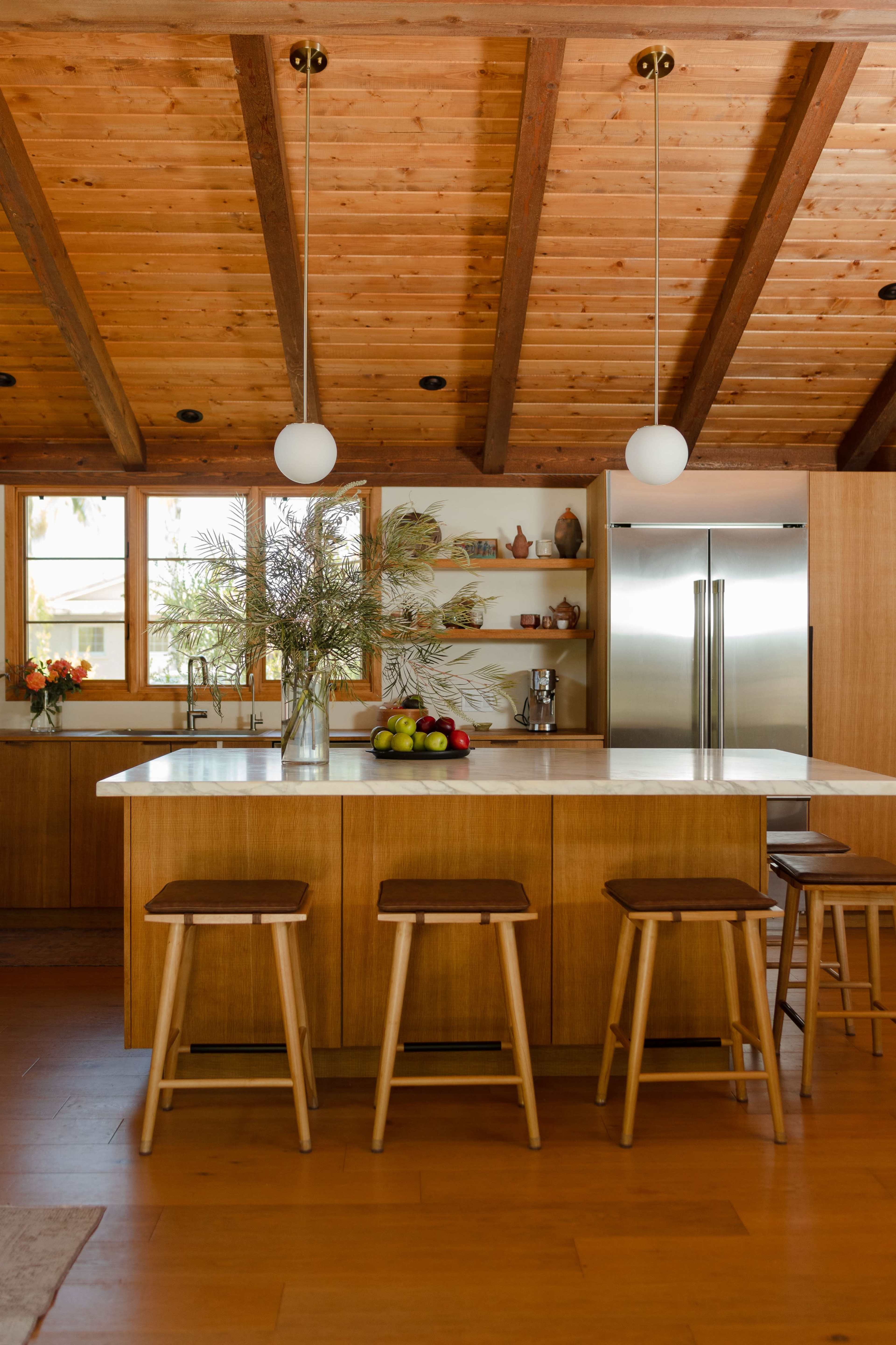 A modern kitchen features wooden beams, ample natural light, and a central island with stools and a vase of dried flowers.