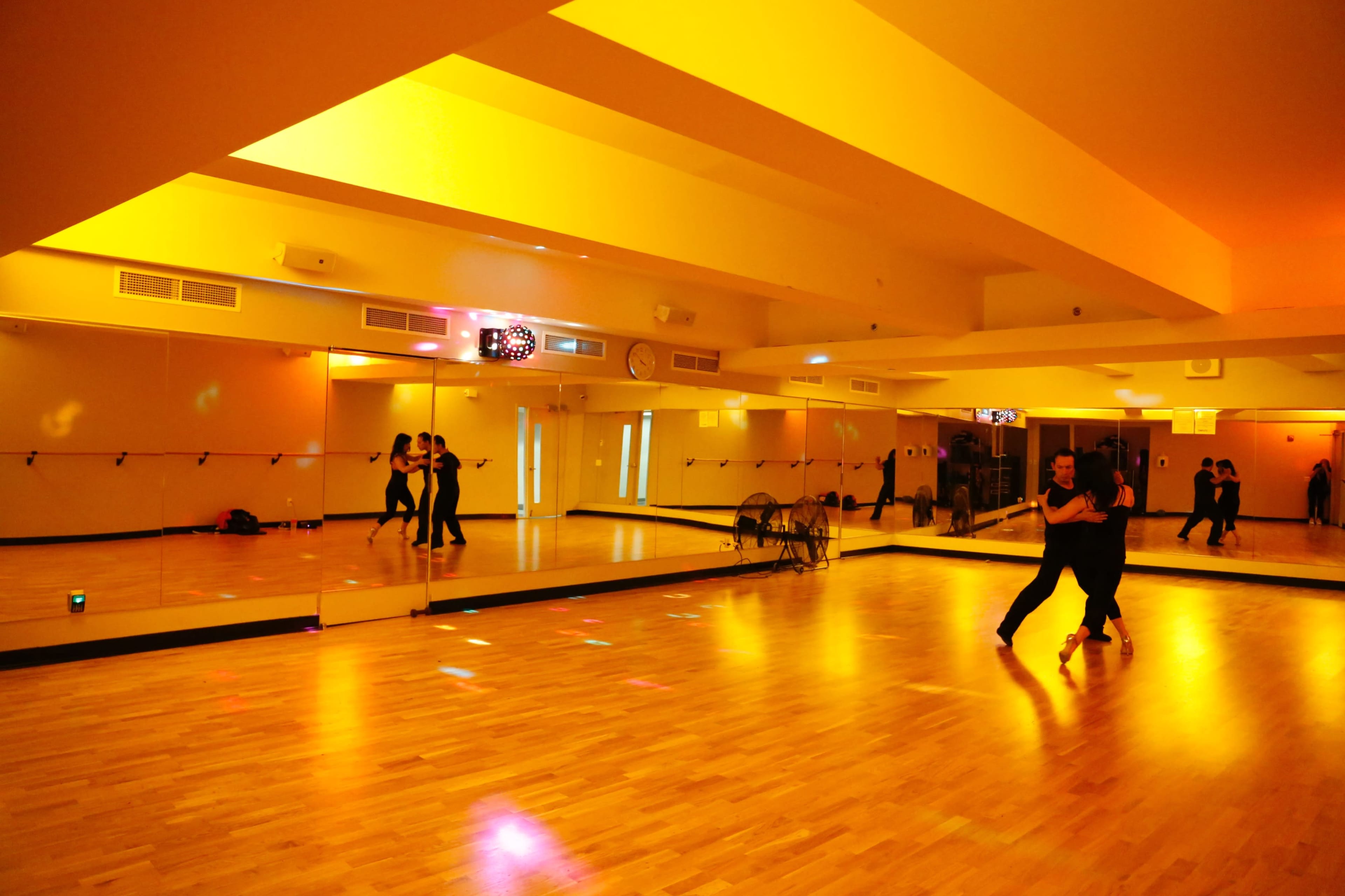 A dance studio with mirrored walls and warm yellow lighting features two dancers practicing in the center of the room.