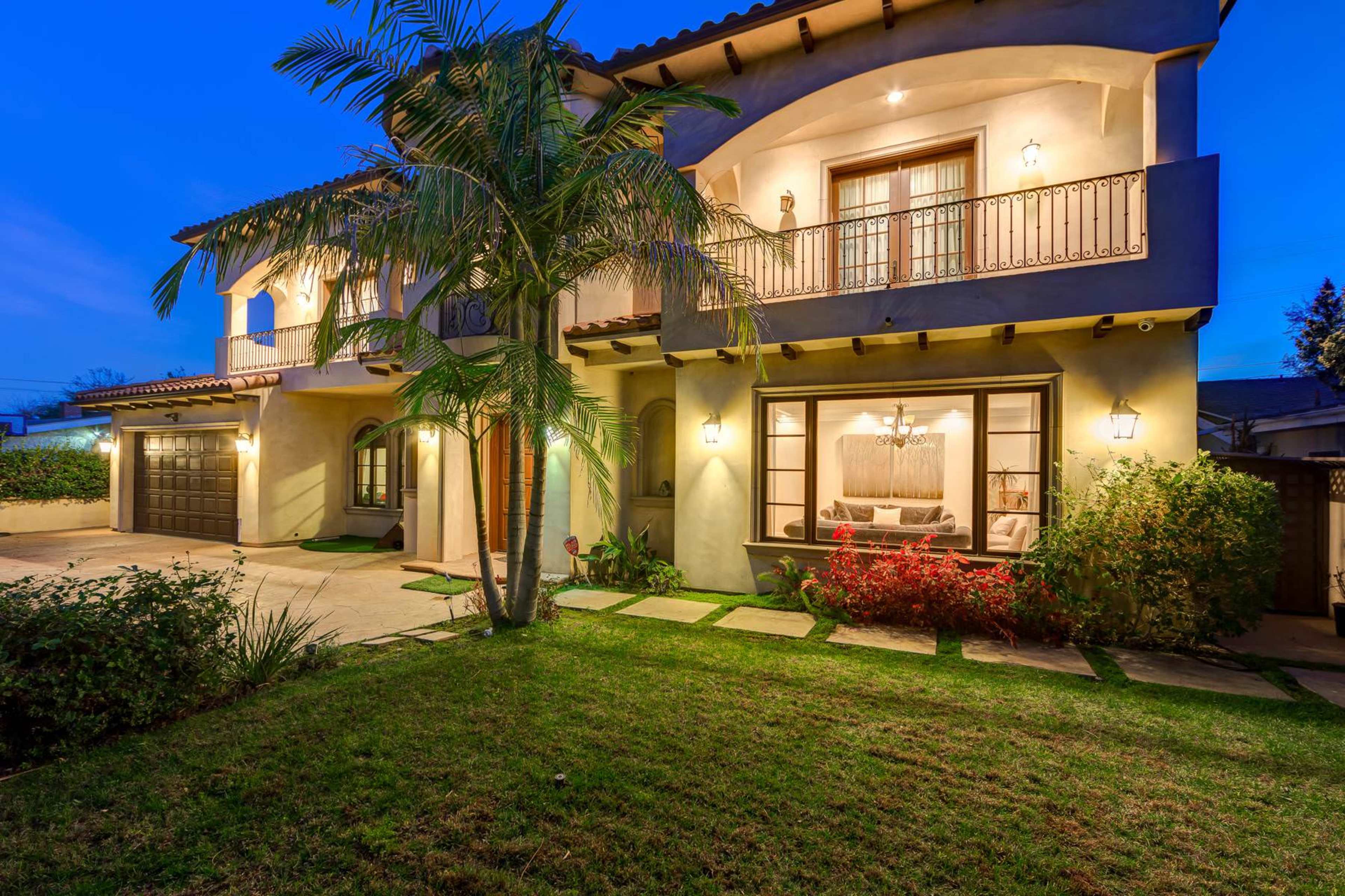 A two-story house with a balcony, surrounded by lush greenery, is illuminated at dusk.