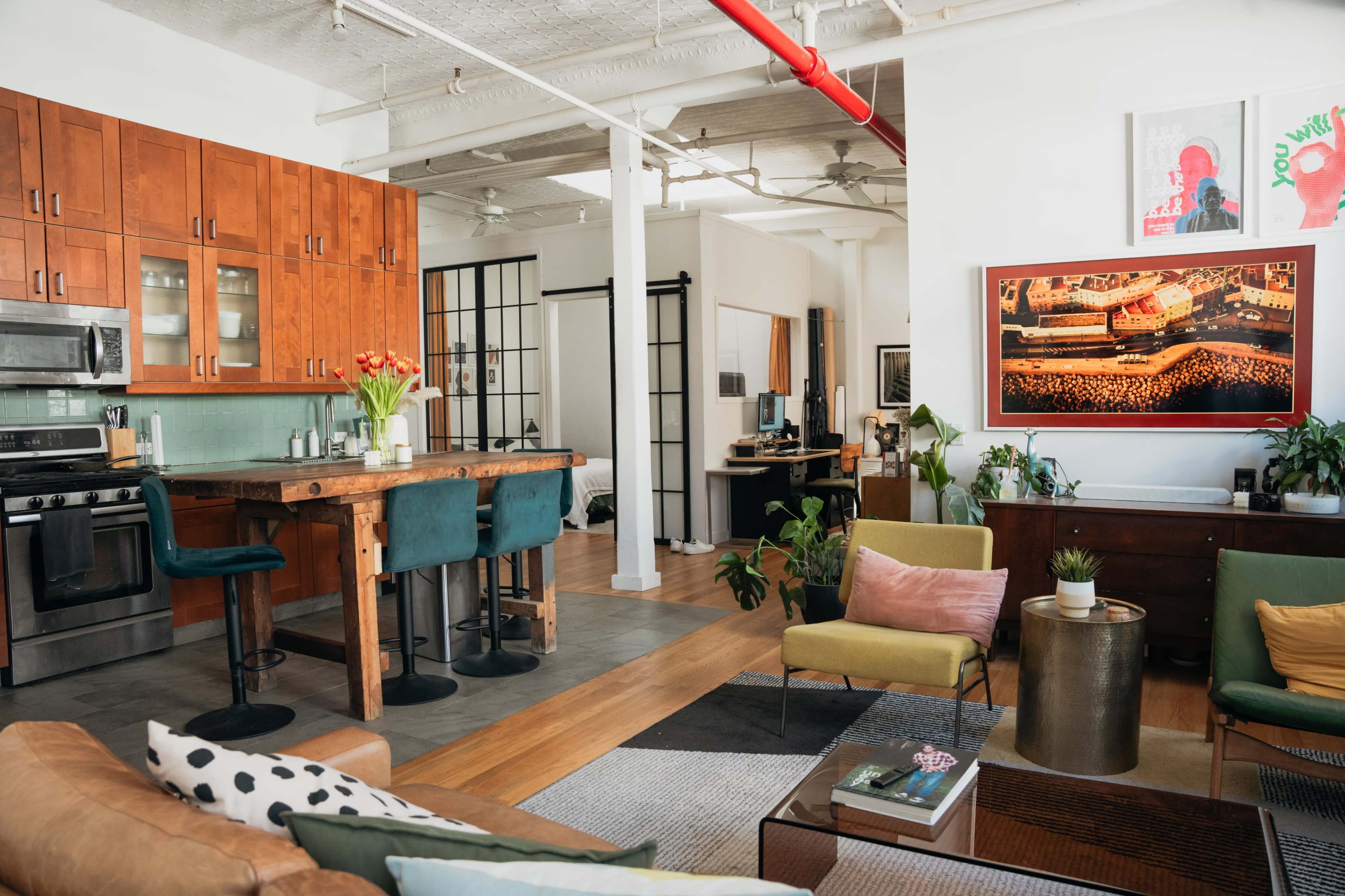 The image shows a modern open-concept living space featuring a kitchen with wooden cabinets, a dining area with bar stools, and a living area furnished with colorful chairs and plants.