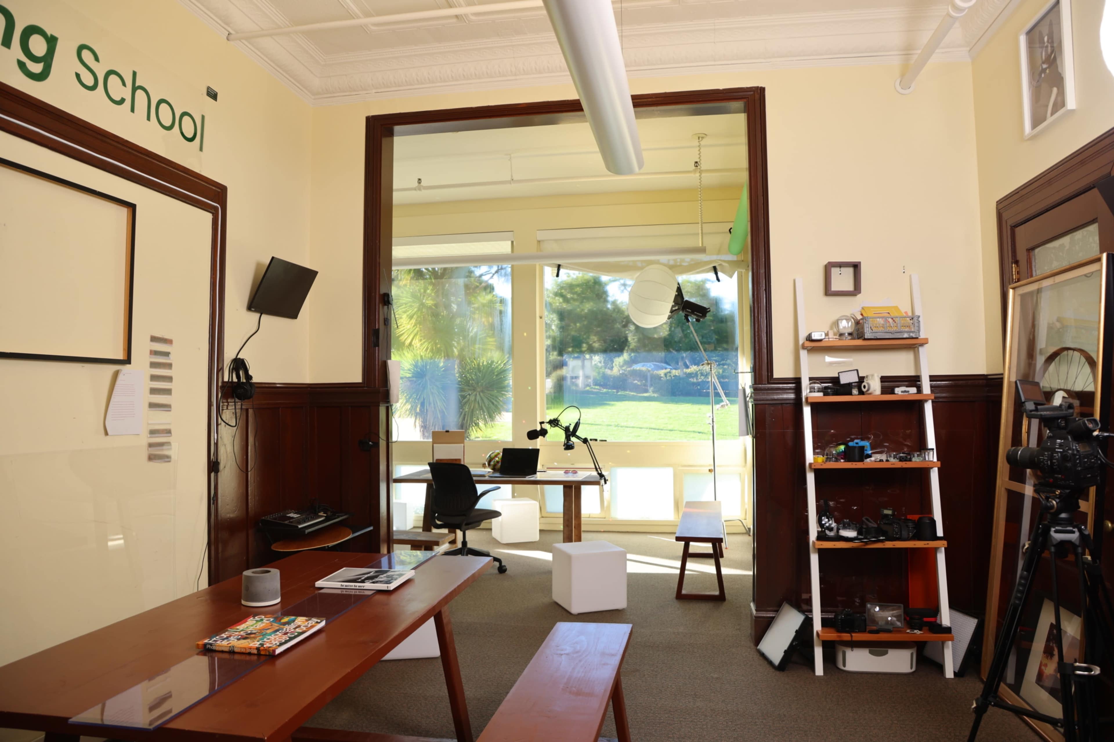 The image shows a classroom with a wooden table, chairs, and a large window overlooking a green area, along with various teaching materials and equipment on shelves.
