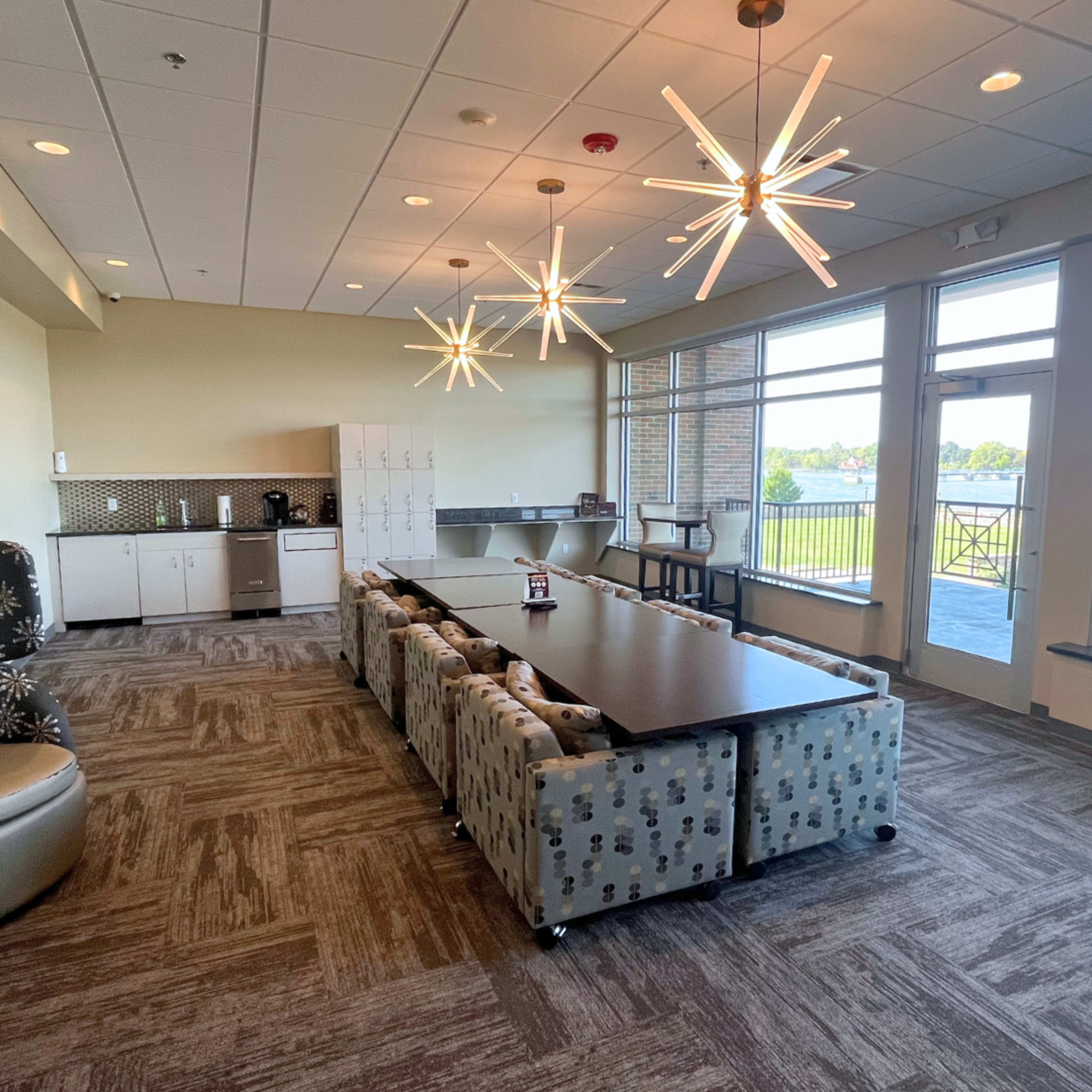 The image shows a modern multi-purpose room with a long table surrounded by patterned chairs, bright lighting fixtures, and windows offering a view of the outdoors.