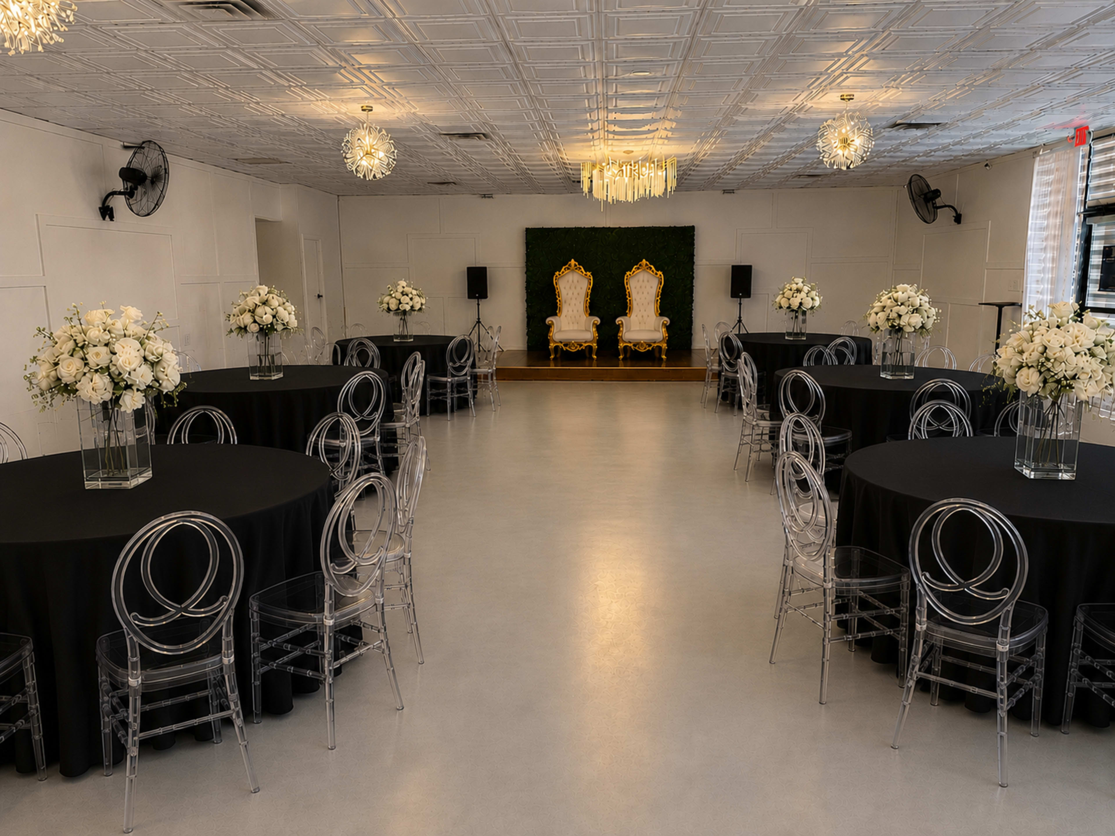 The image shows a spacious event room with two ornate chairs positioned against a green backdrop, surrounded by elegantly set tables with black tablecloths and clear acrylic chairs.