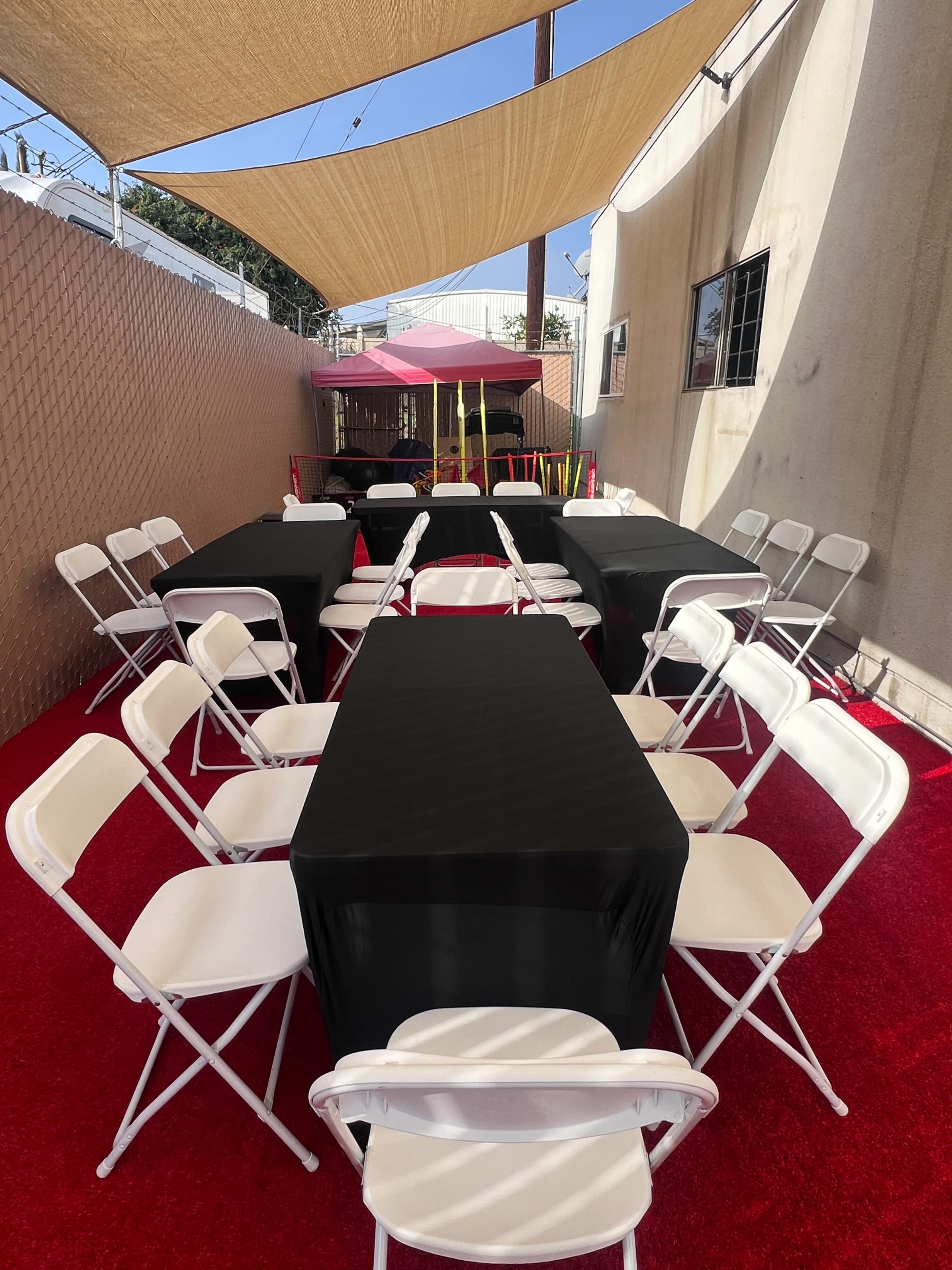 The image shows a series of black-covered tables set up with white folding chairs under a shaded area.