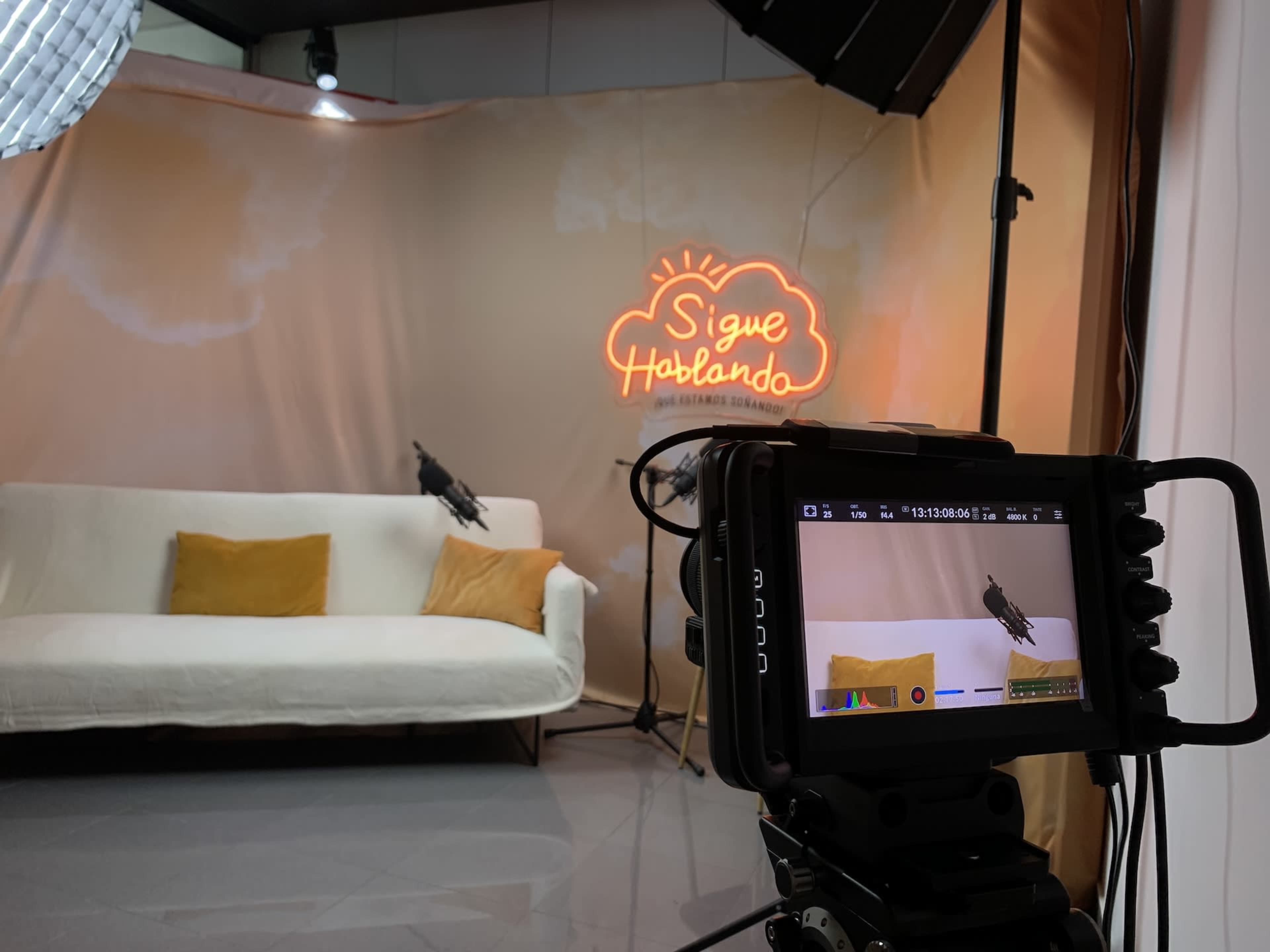A camera setup is focused on a white sofa with yellow cushions in front of a backdrop featuring a neon sign that reads "Sigue Hablando."