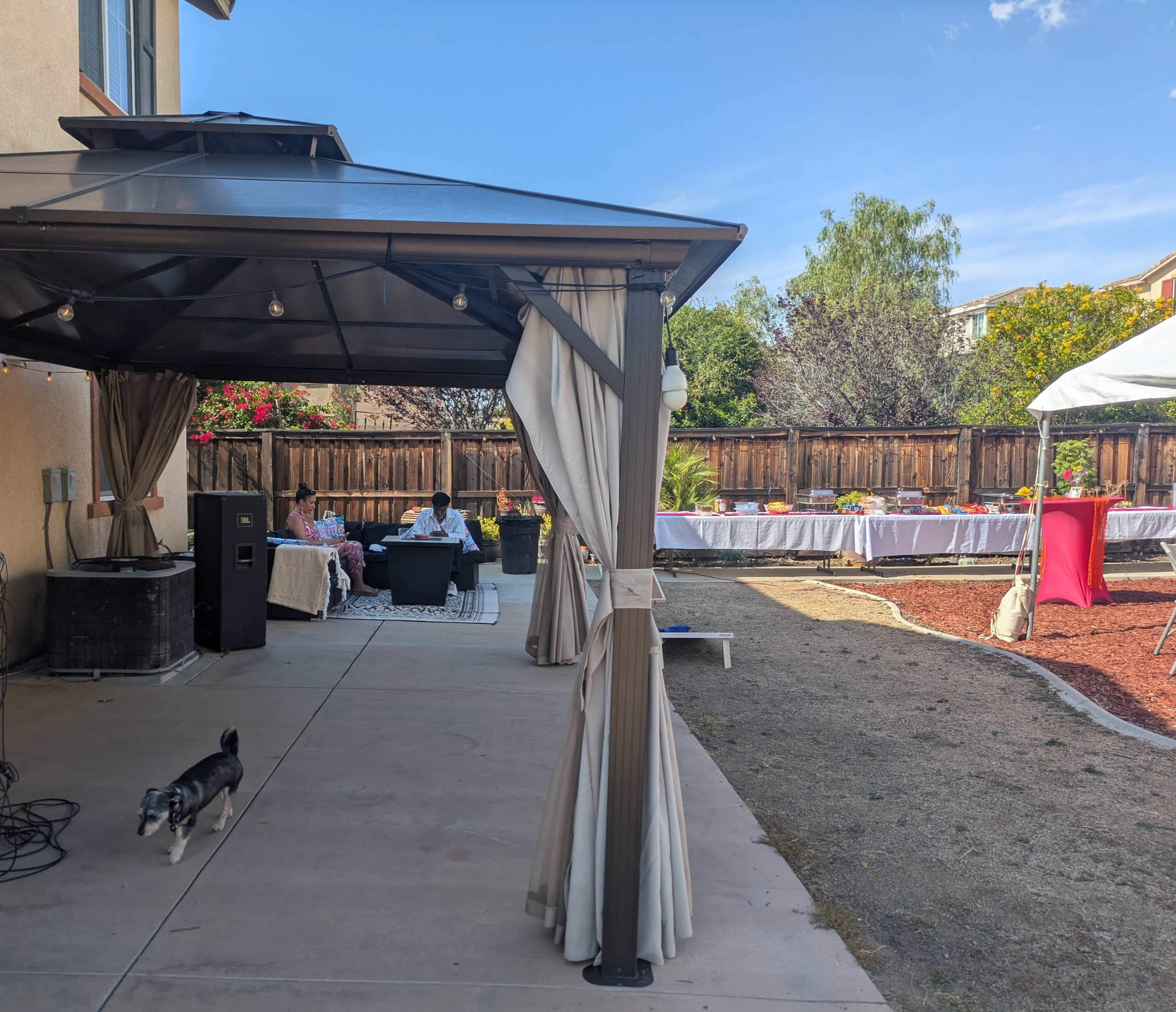 A backyard setting with a gazebo, seating area, and a party setup featuring tables and decorations, along with a small dog in the foreground.