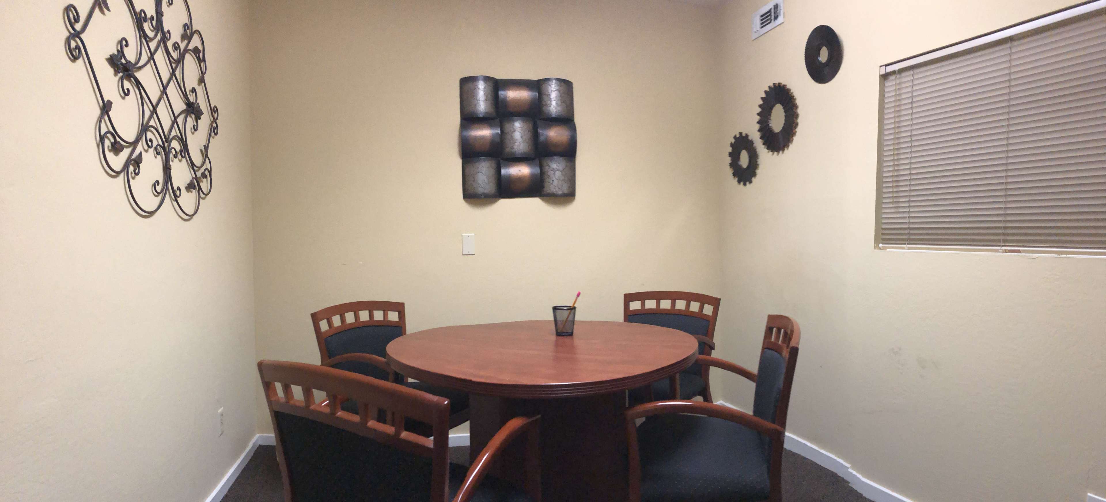 A small room features a round wooden table surrounded by four chairs, with decorative wall art and a window on one side.