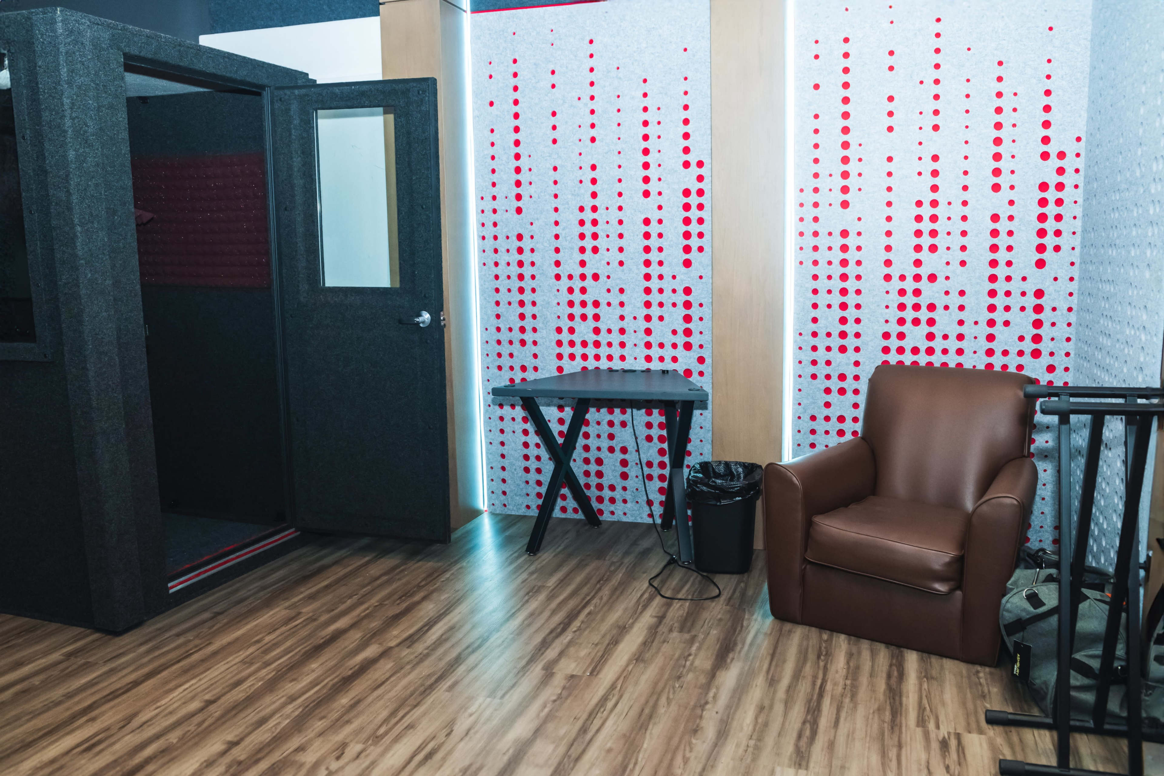 The image shows a room with a sound booth, a brown leather chair, a small table, and a patterned wall featuring red dots.