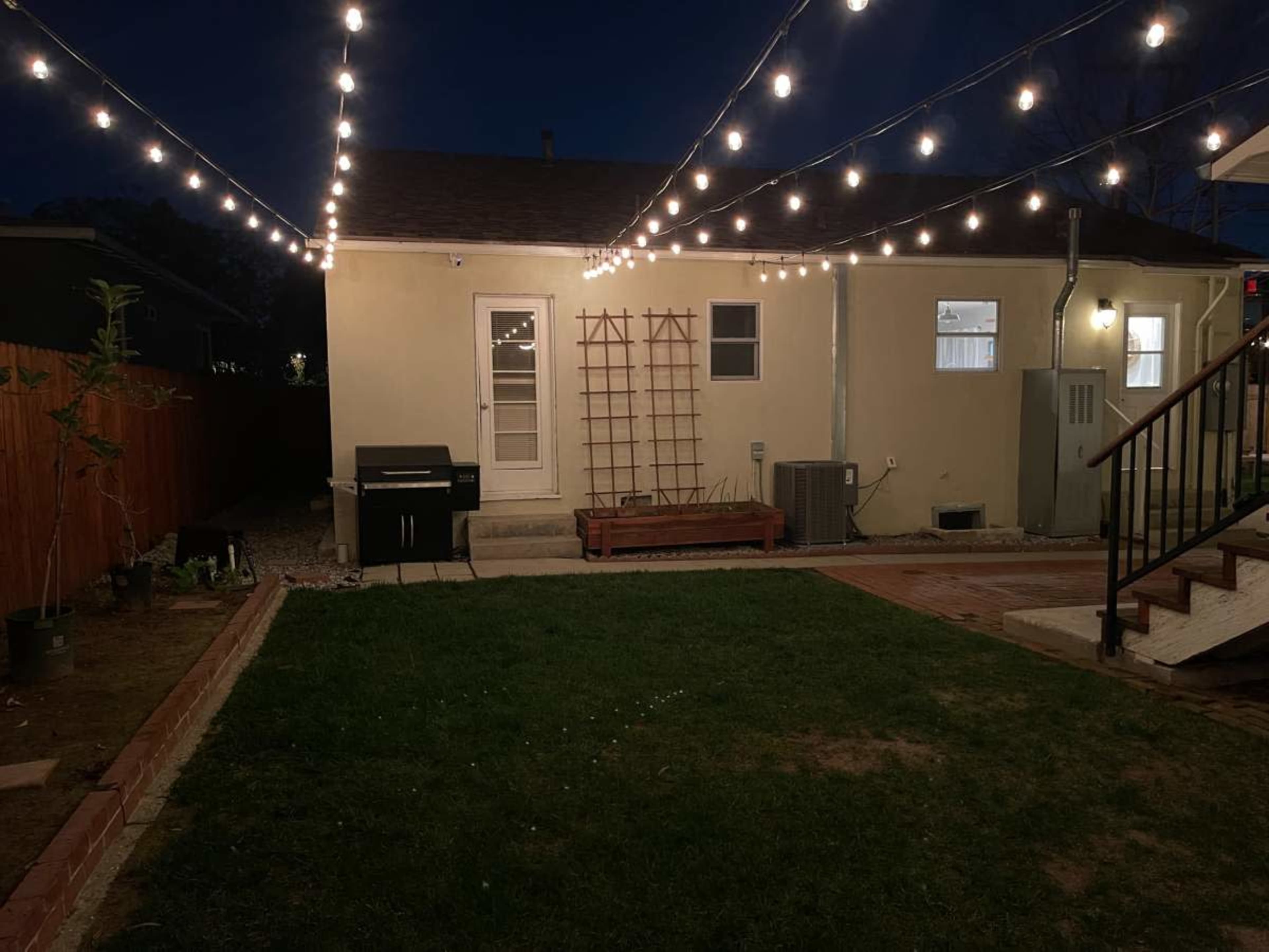 A backyard at night with string lights overhead, a grill, a wooden bench, and a small trellis against the wall of a house.