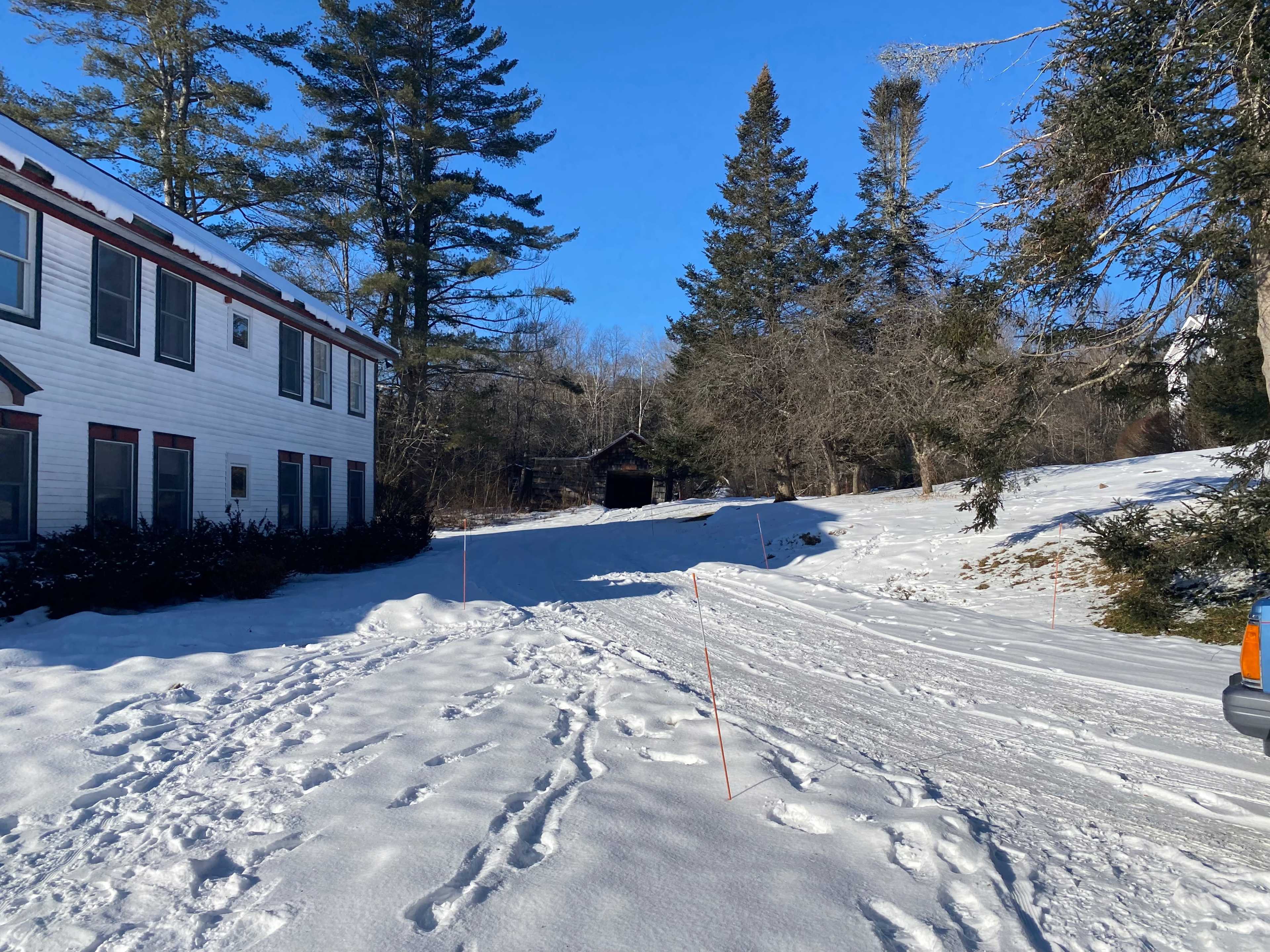 A snow-covered path leads to a dark structure surrounded by trees beside a two-story building.