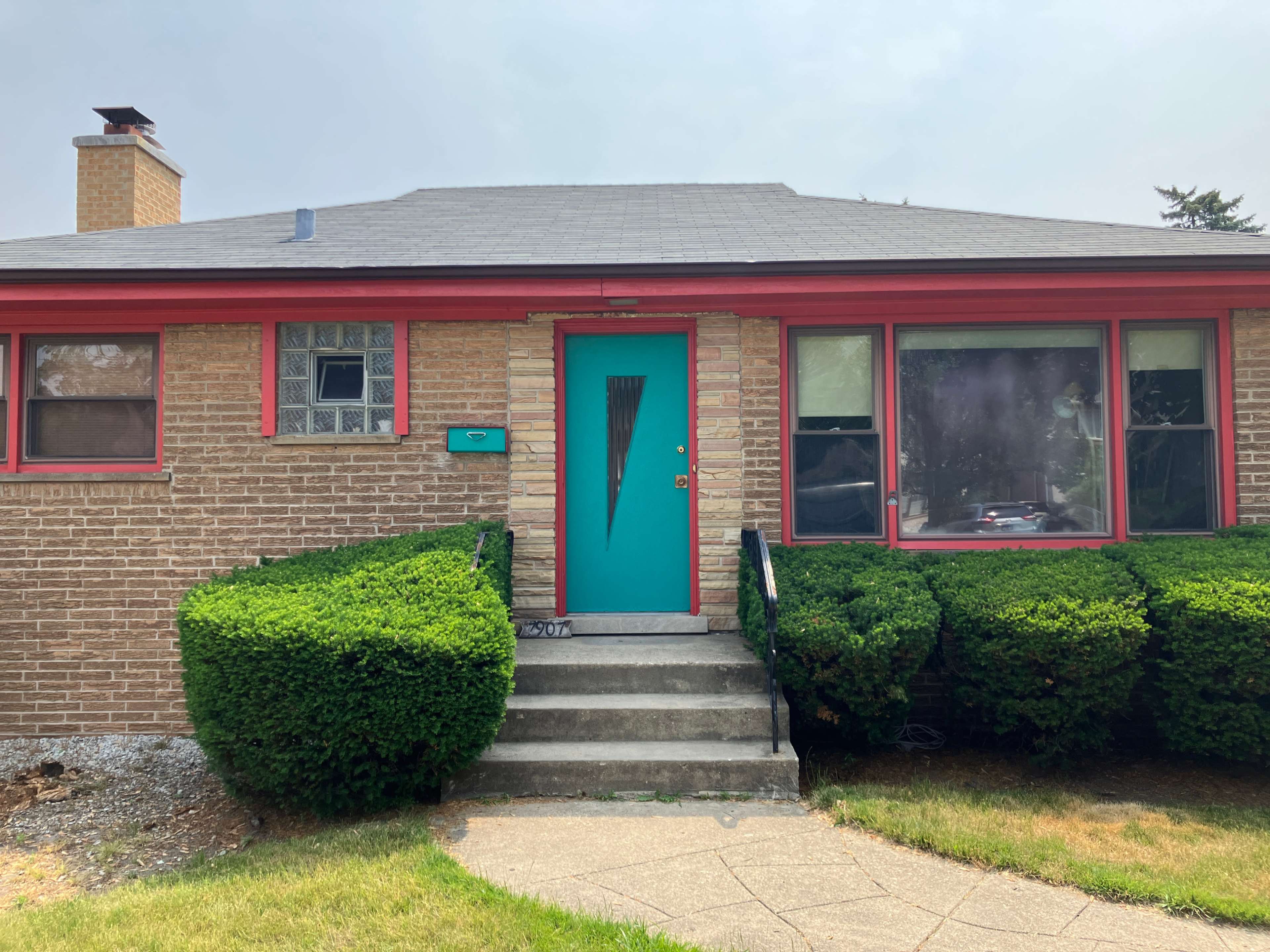 The image shows a bungalow-style house with a turquoise front door and vibrant red trim, surrounded by neatly trimmed bushes and a concrete pathway.