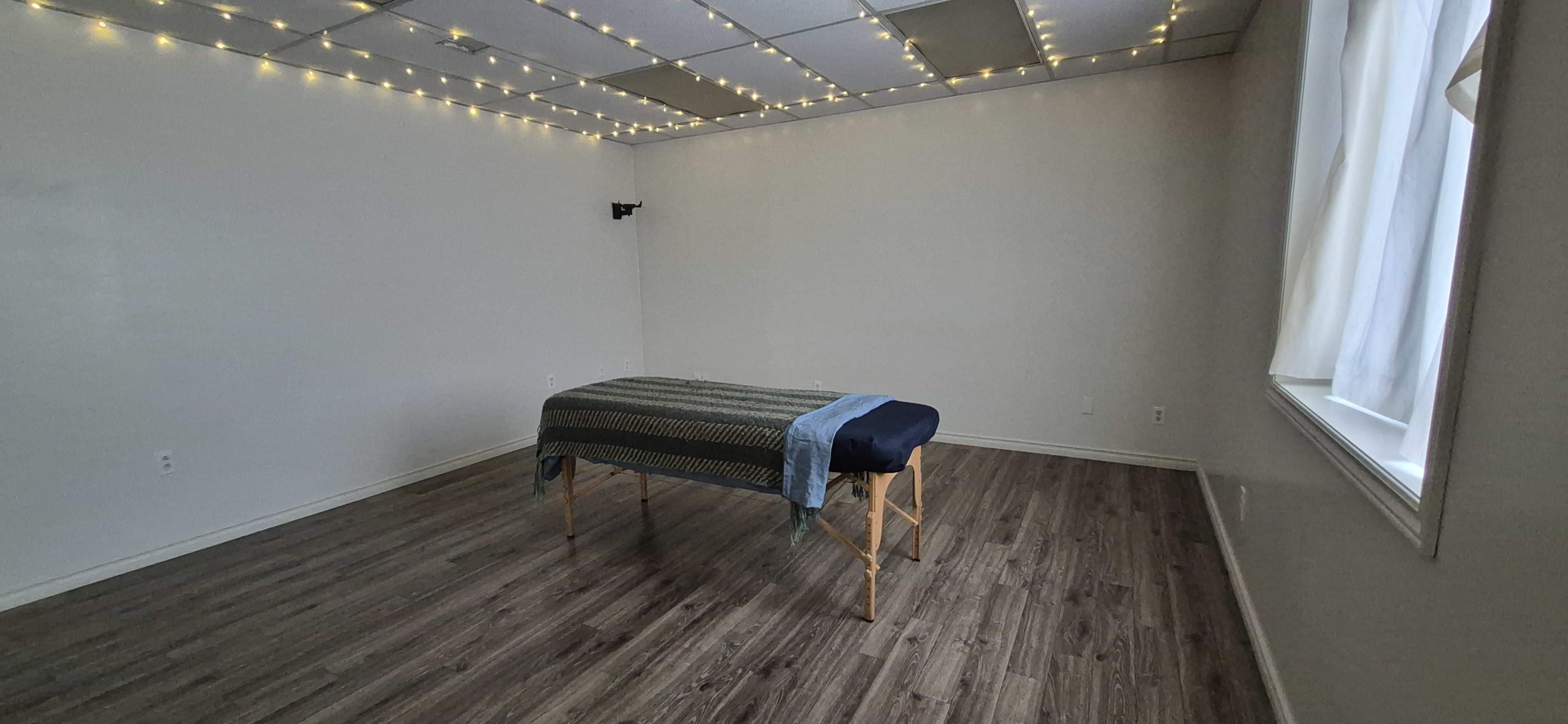 A massage table covered with a blue sheet is positioned in a sparsely decorated room with string lights along the ceiling.