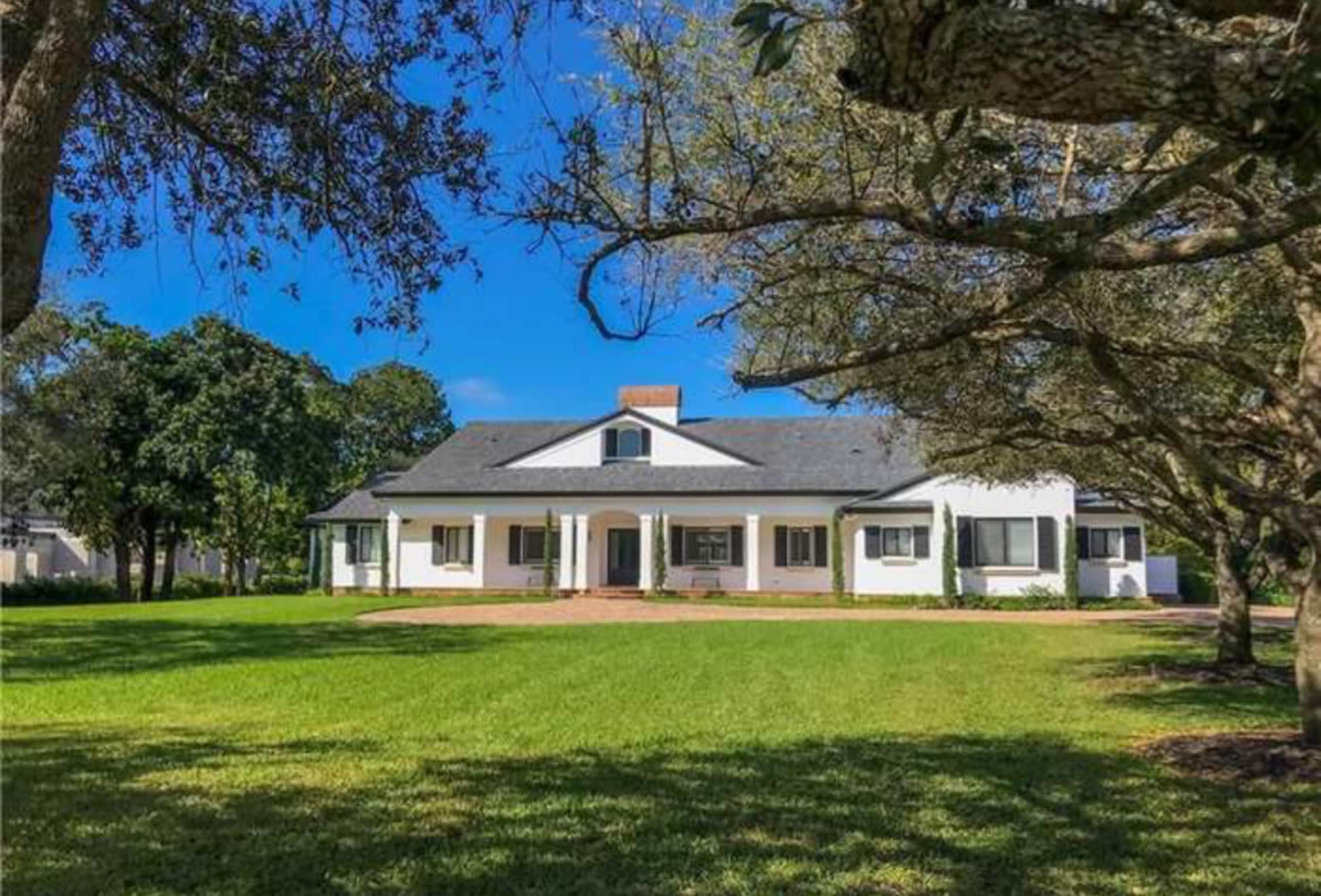 A single-story white house with a black roof and a central chimney sits on a green lawn surrounded by trees.
