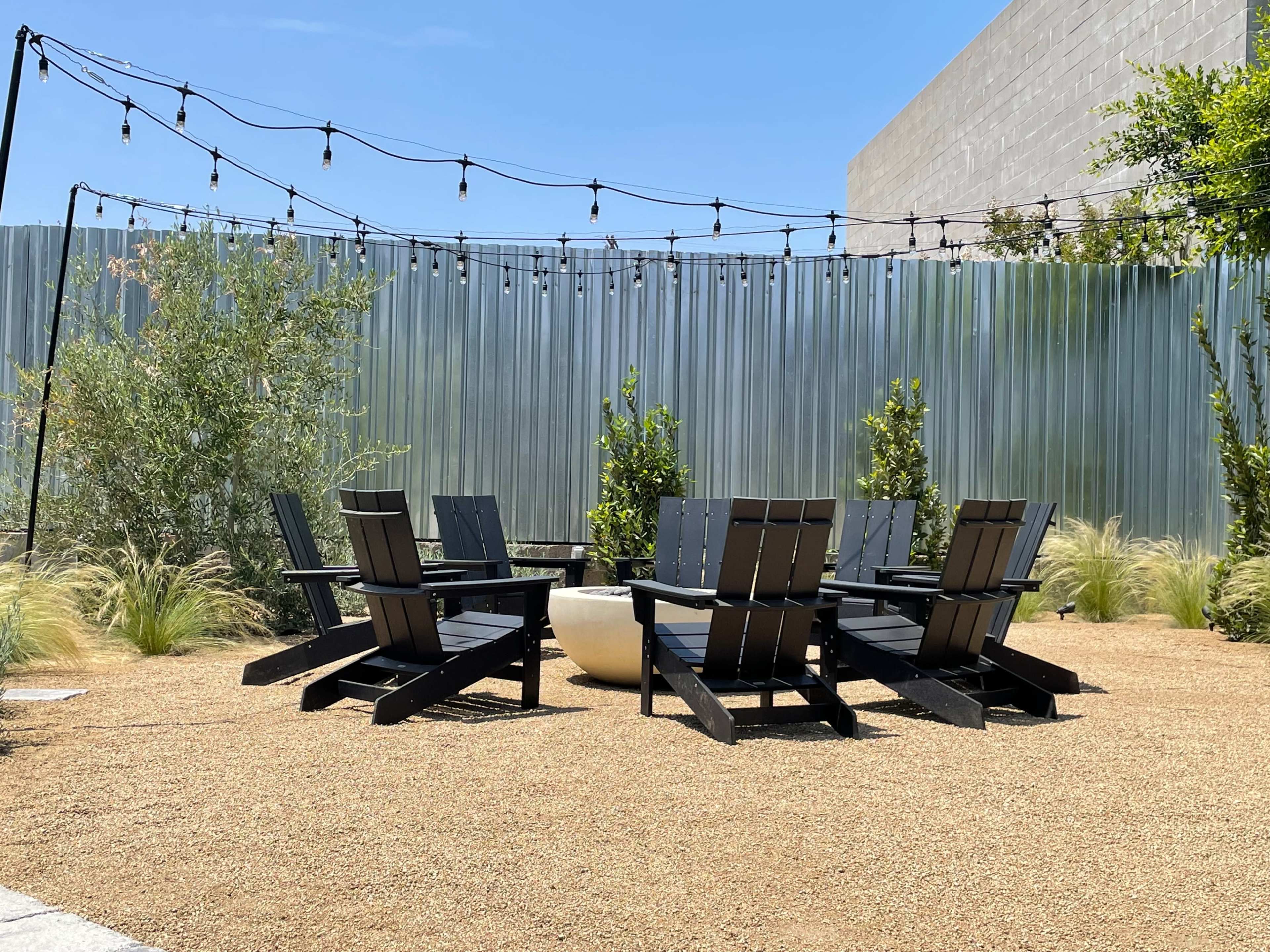 The image shows a circular arrangement of black Adirondack chairs surrounding a central fire pit, set on gravel with green plants and string lights overhead.