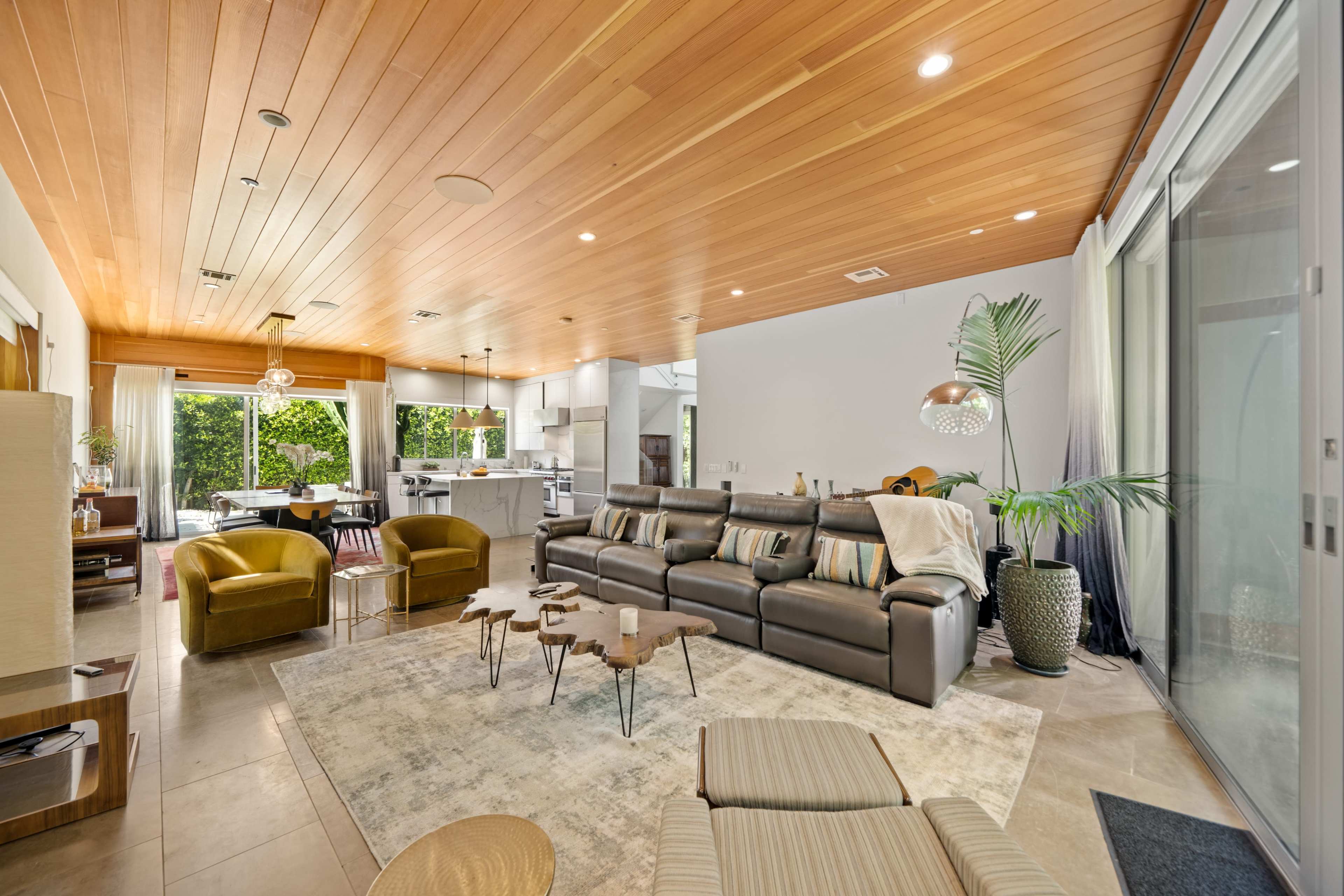 The spacious living room features a brown leather sectional sofa, two green armchairs, a large rug, and large windows overlooking a green area.