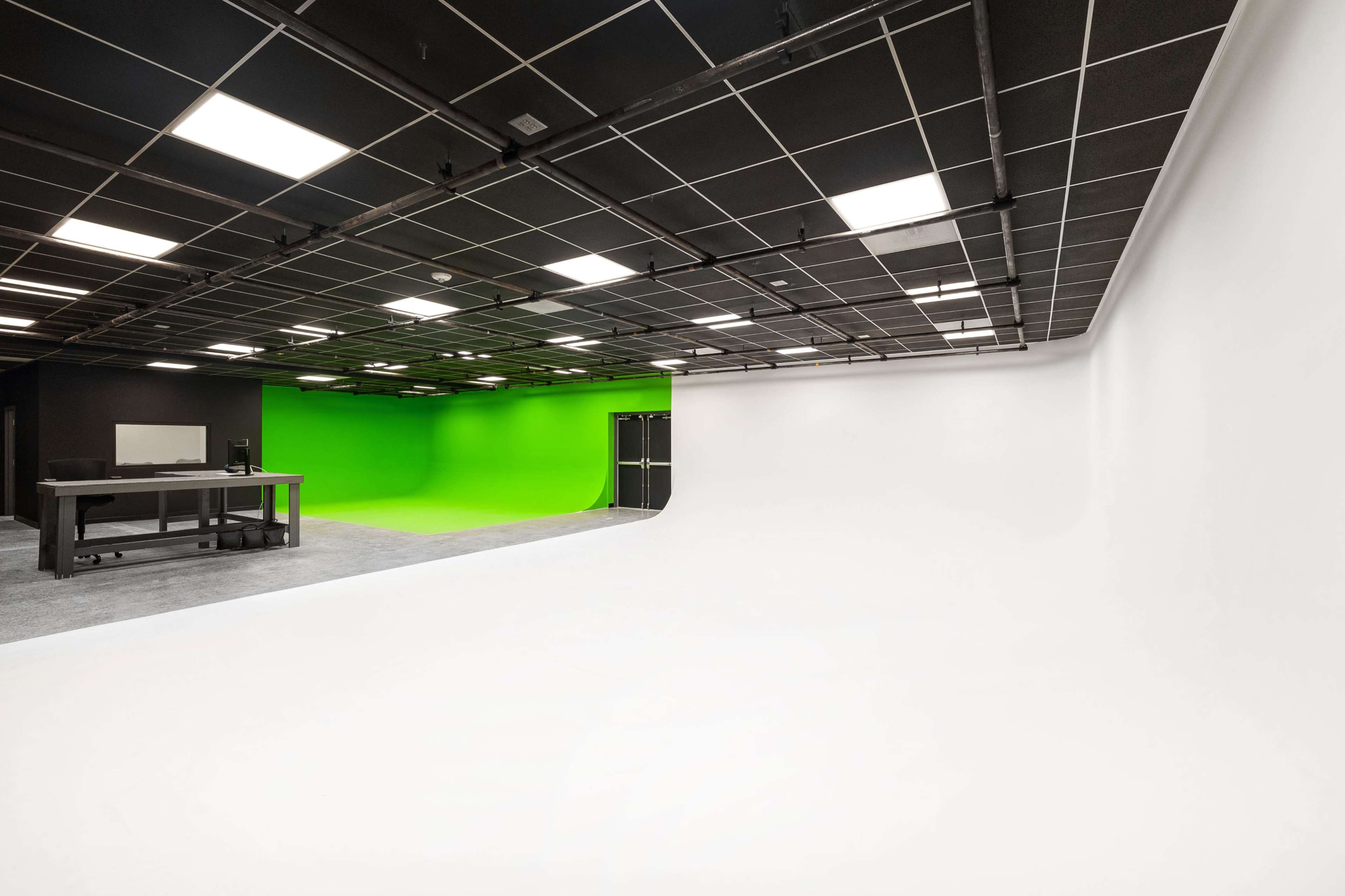 A spacious studio features a white background on one side and a green screen on the other, with a black ceiling and overhead lights.