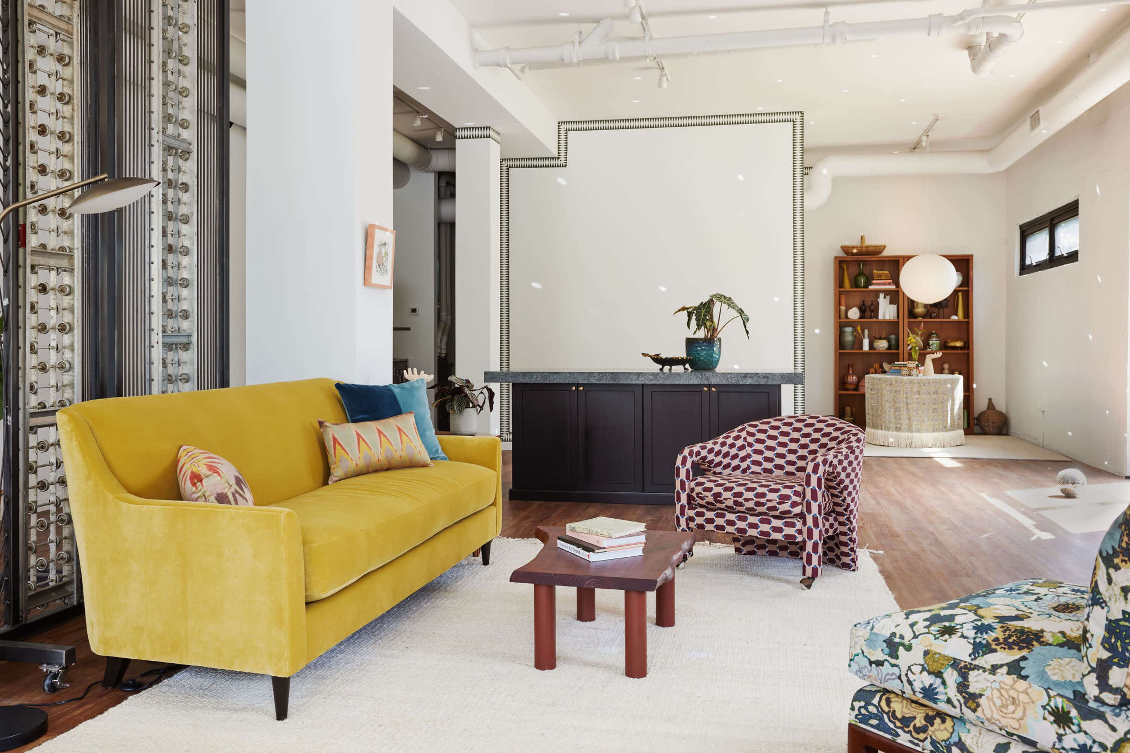 A bright living room features a yellow sofa, patterned armchair, wooden coffee table, and a large white rug.