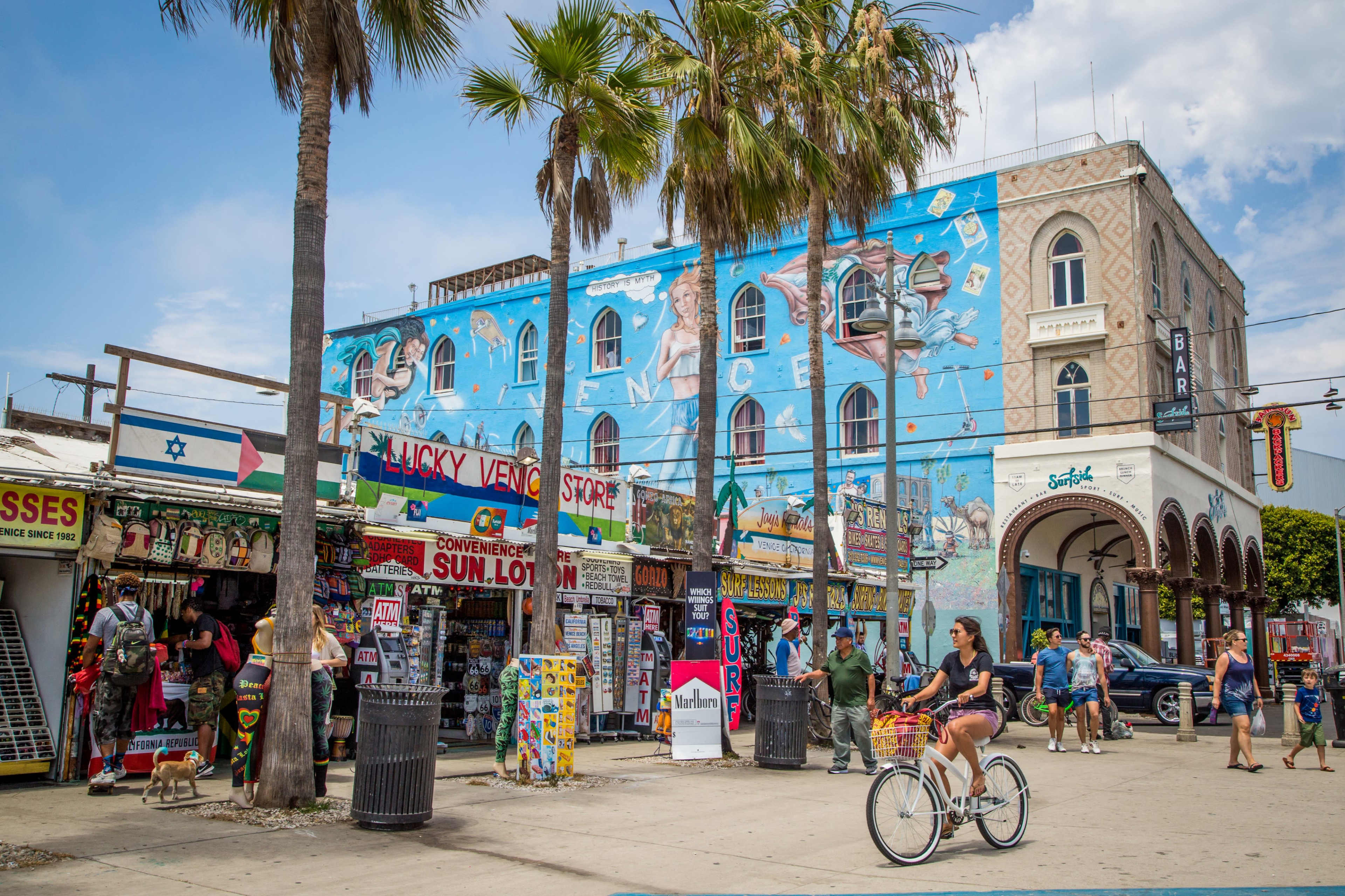 A person rides a bicycle past colorful shops and a mural on a building in a busy outdoor area.