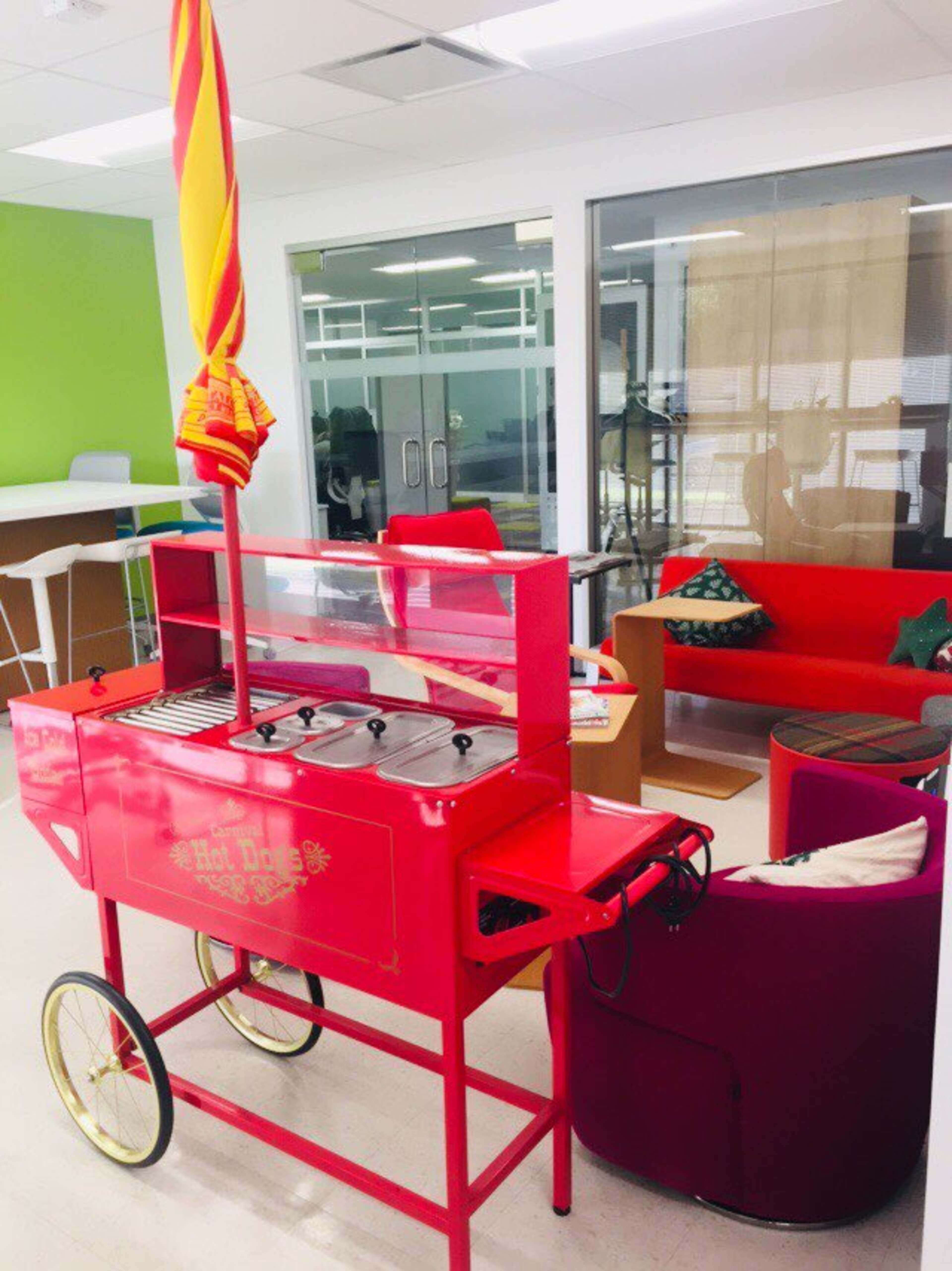 A red hot dog cart with a striped umbrella stands in a modern office space featuring colorful furniture.
