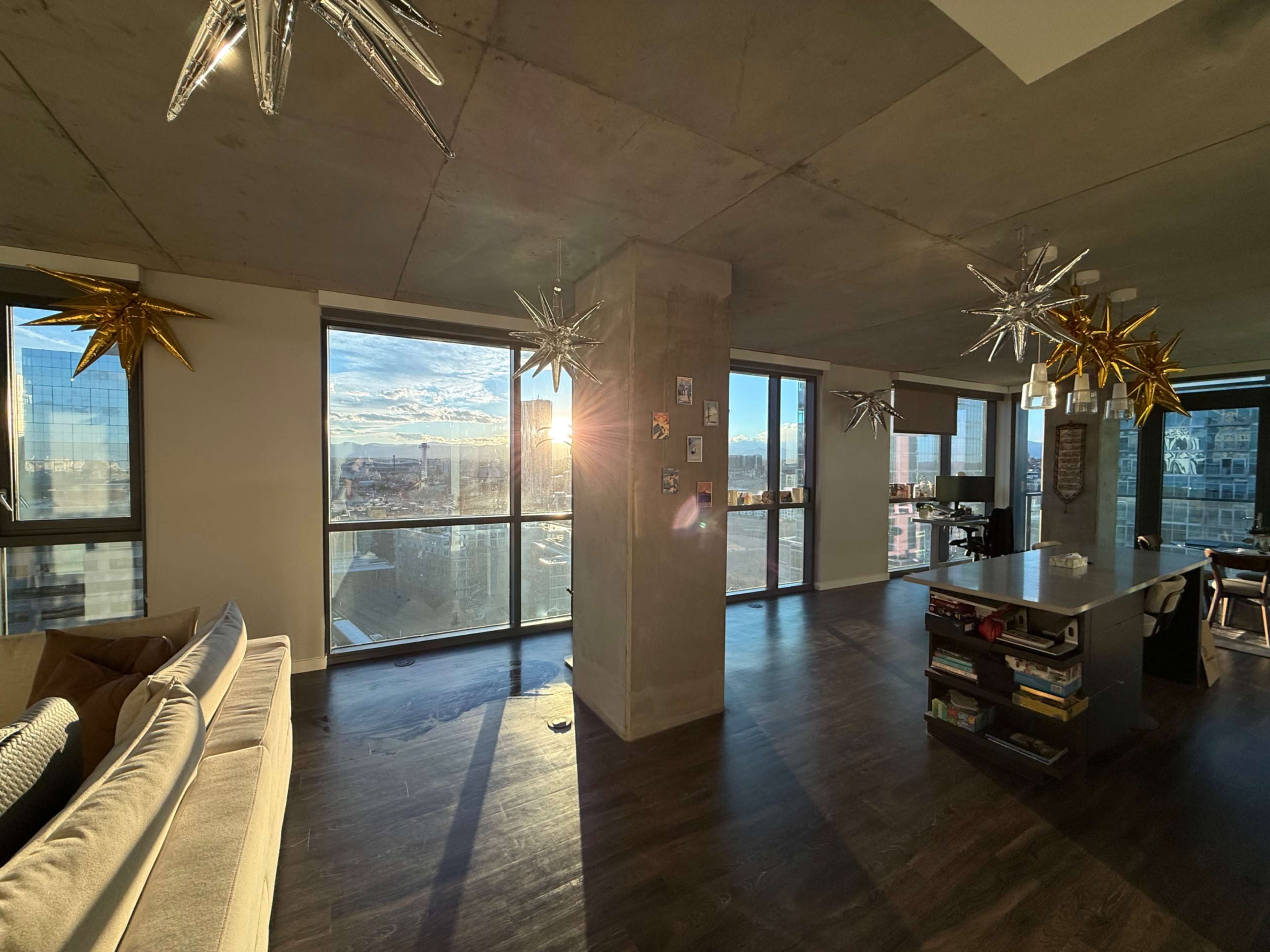 The interior of a modern apartment features large windows with a sunset view, hardwood flooring, and stylish star-shaped pendant lights hanging from the ceiling.