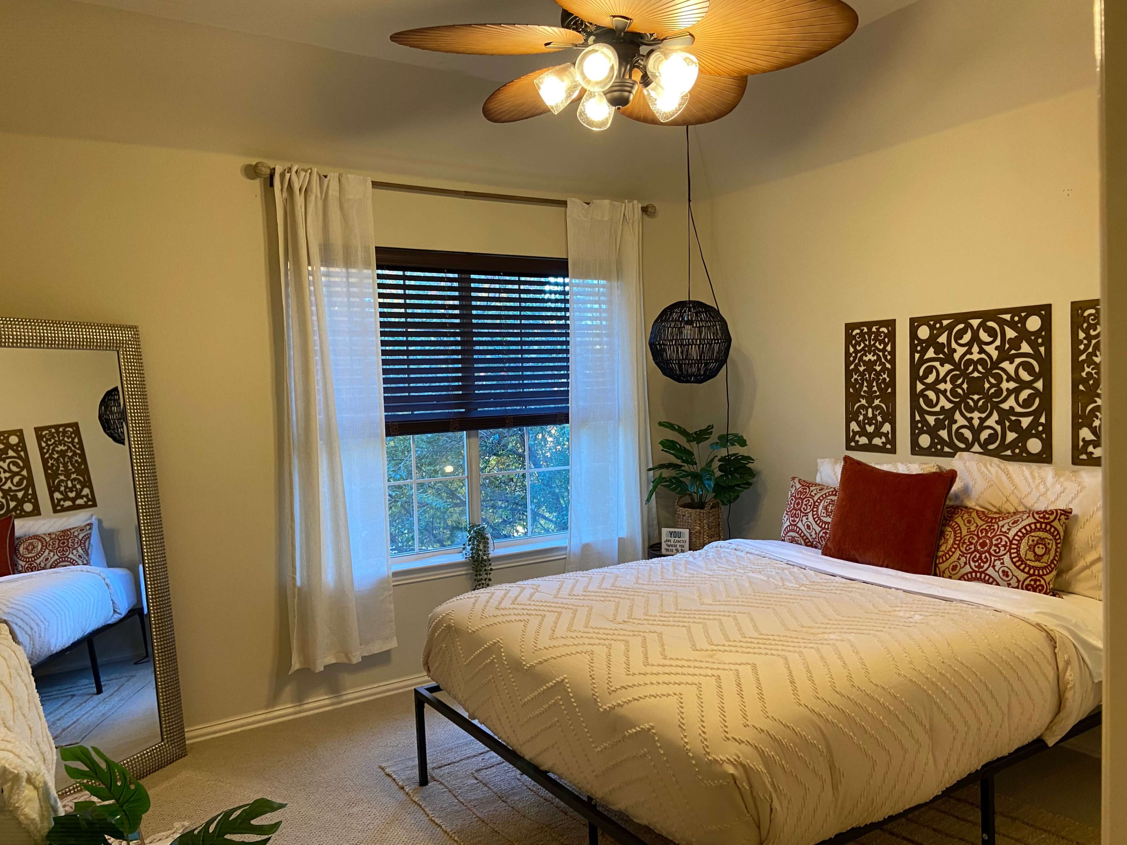 A well-decorated bedroom features a bed with patterned bedding, a large mirror, and a window with venetian blinds, complemented by plants and a ceiling fan.