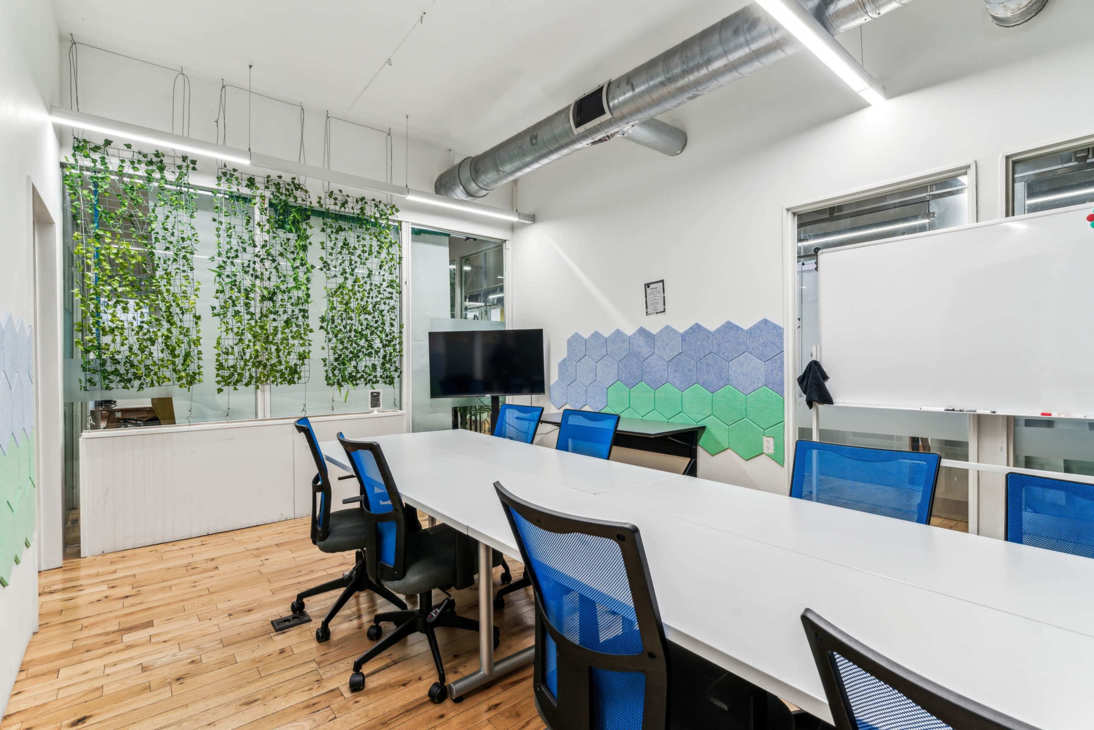 The image shows a modern conference room featuring a long white table surrounded by blue office chairs, with large windows and greenery decor on the walls.