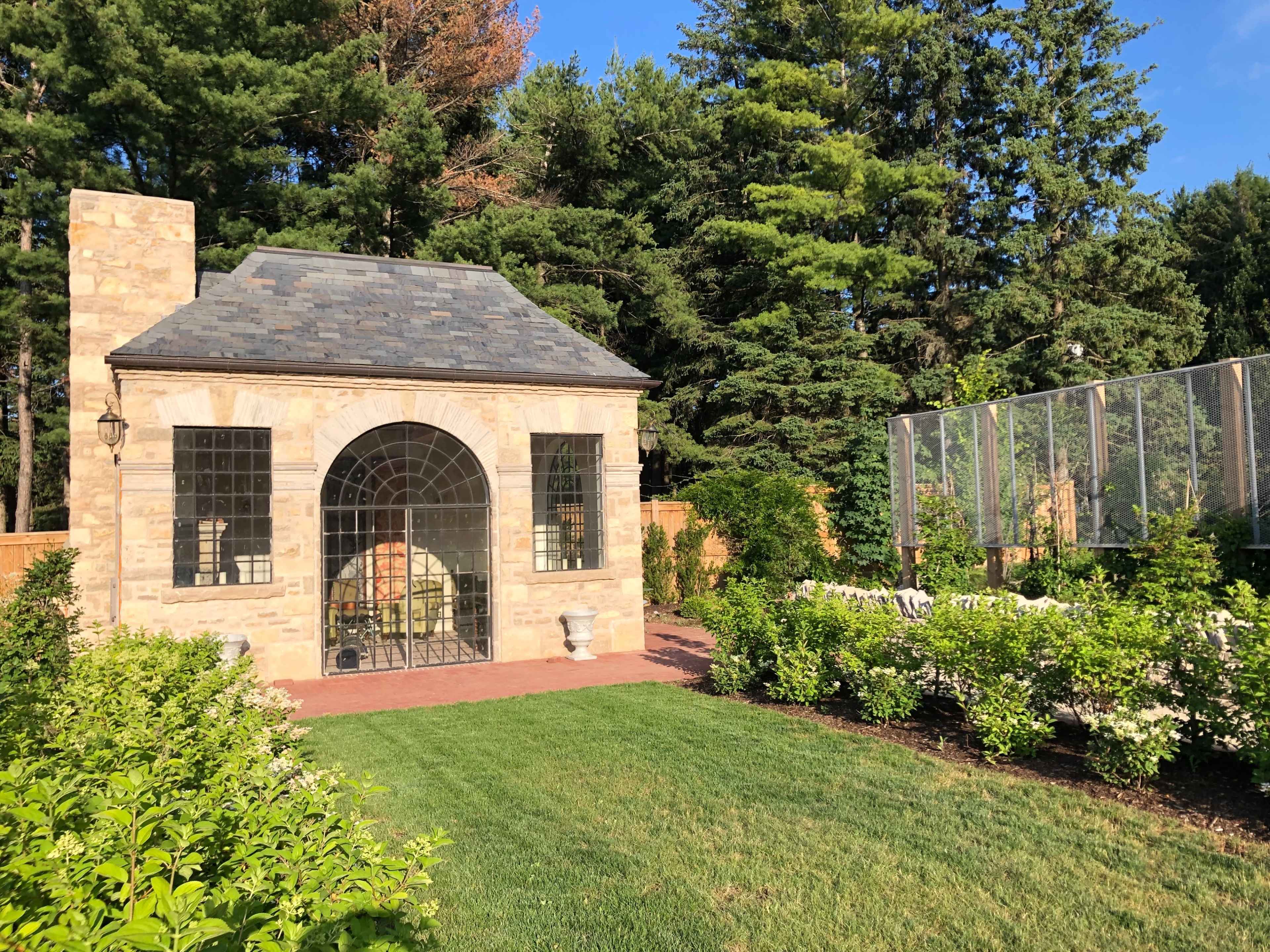 A stone garden structure with a large arched window surrounded by manicured greenery and trees.