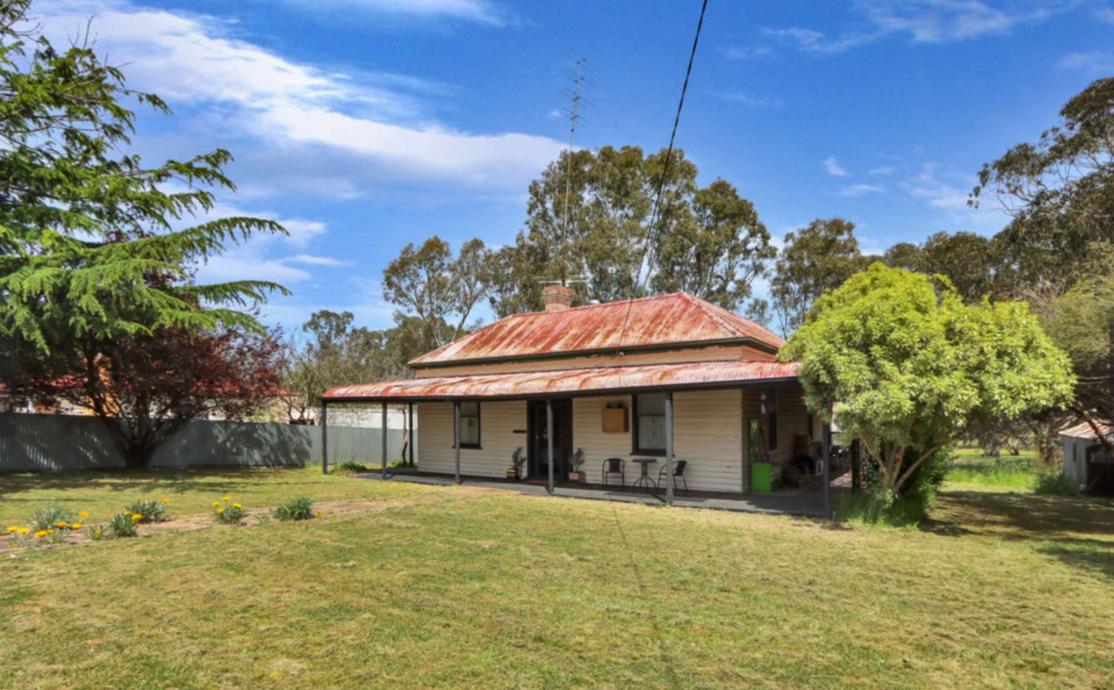 A single-story farmhouse with a corrugated metal roof is set in a grassy yard surrounded by trees and flower beds.