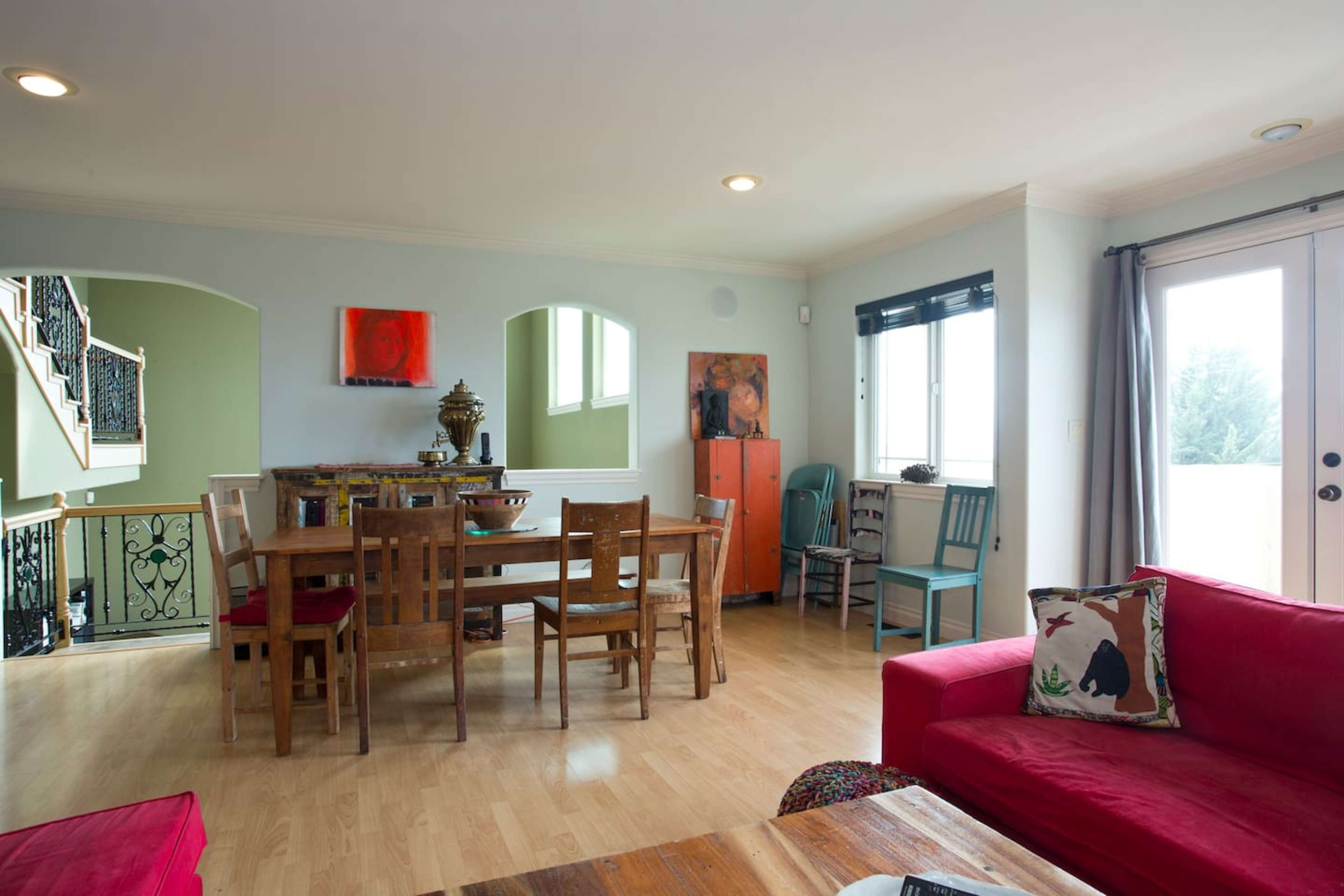 The room features a dining table surrounded by wooden chairs, with a red sofa in the foreground and large windows letting in natural light.