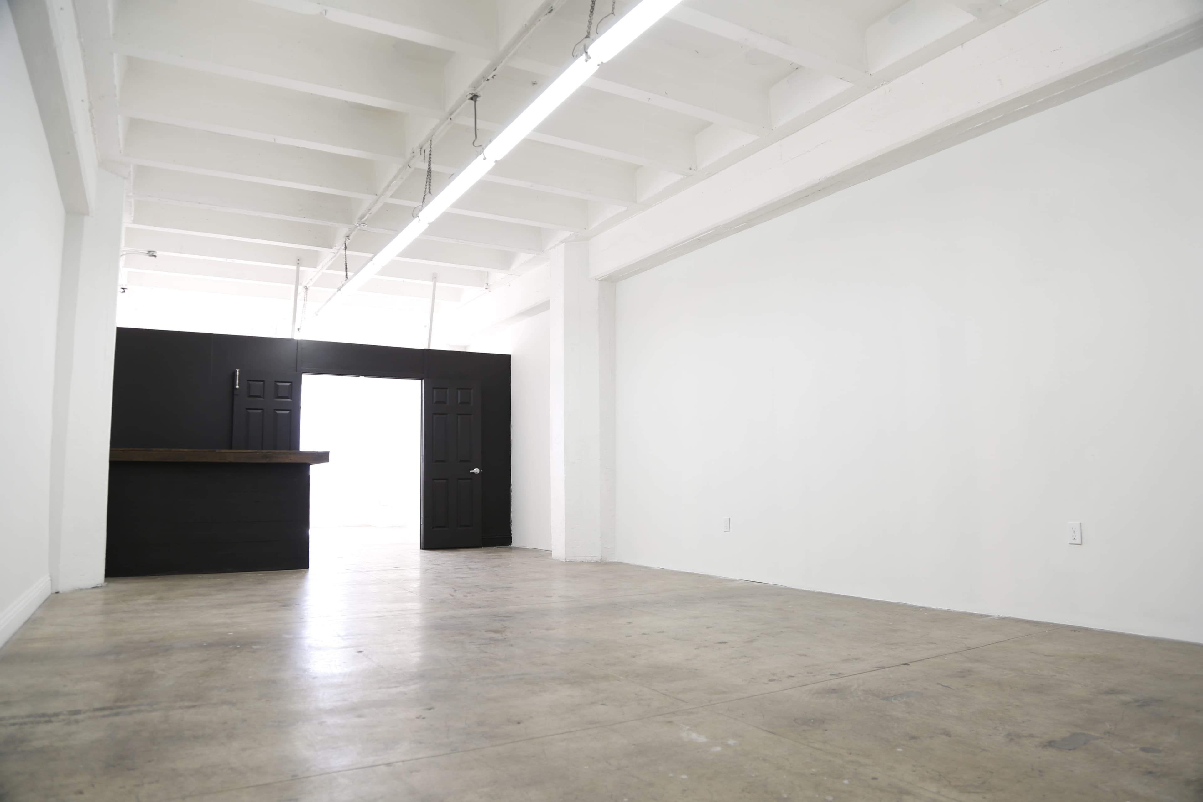 A minimalist interior space with a polished concrete floor, white walls, and a dark wooden counter against a black wall.