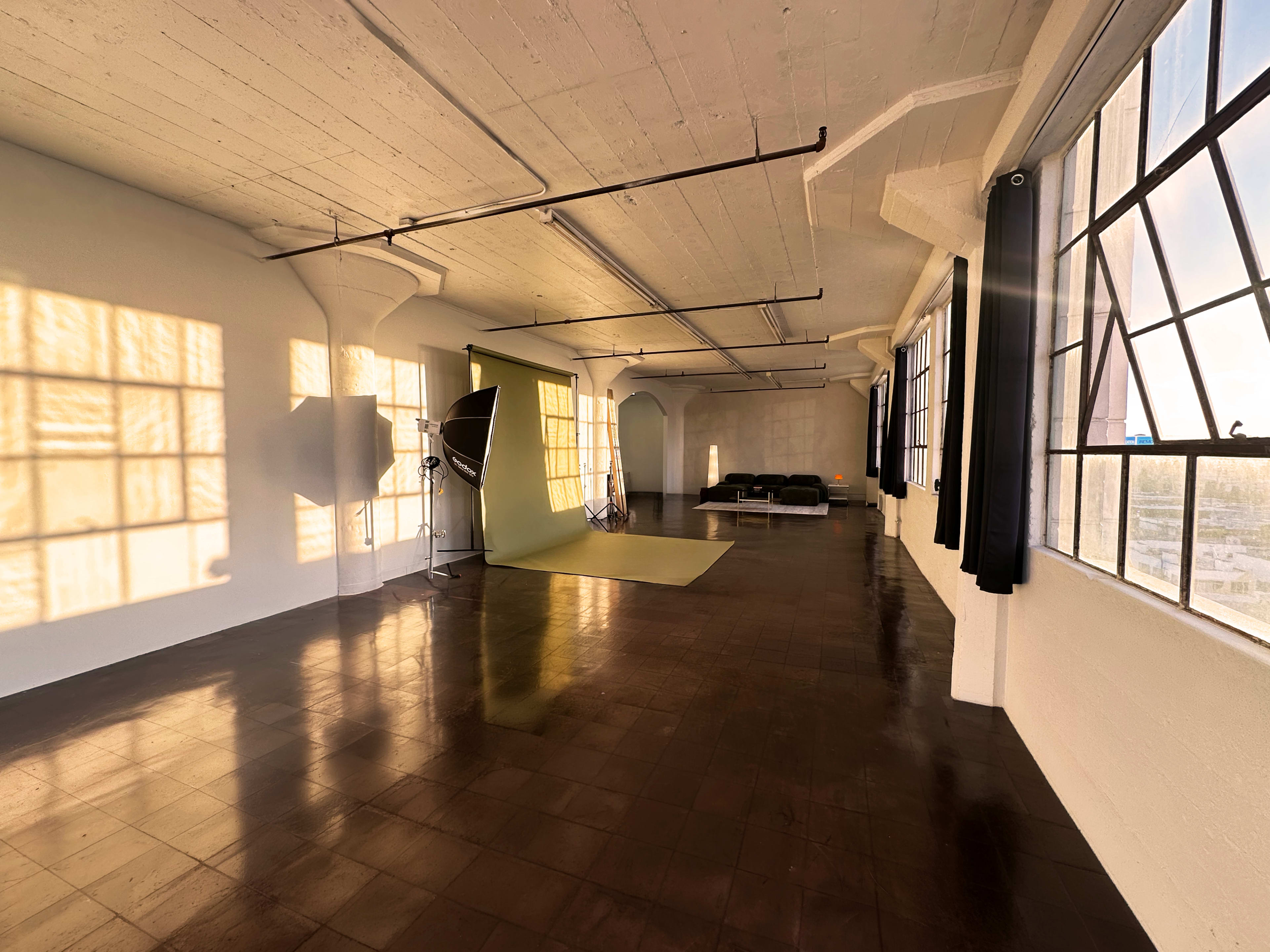 The image shows a spacious interior with large windows casting shadows on the polished floor, featuring a green backdrop, photography equipment, and a black couch.