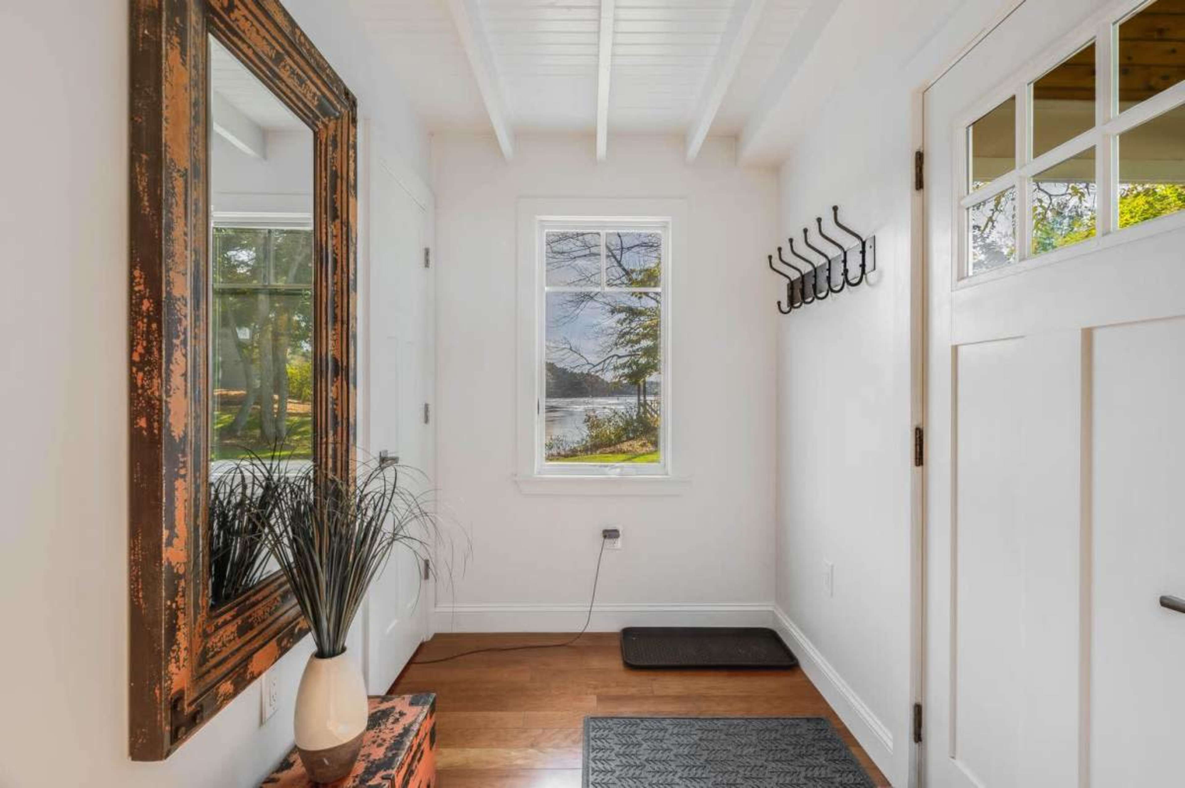 The image shows a light-filled entryway with a wooden mirror, a window overlooking a landscape, a small coat rack, and a console table.