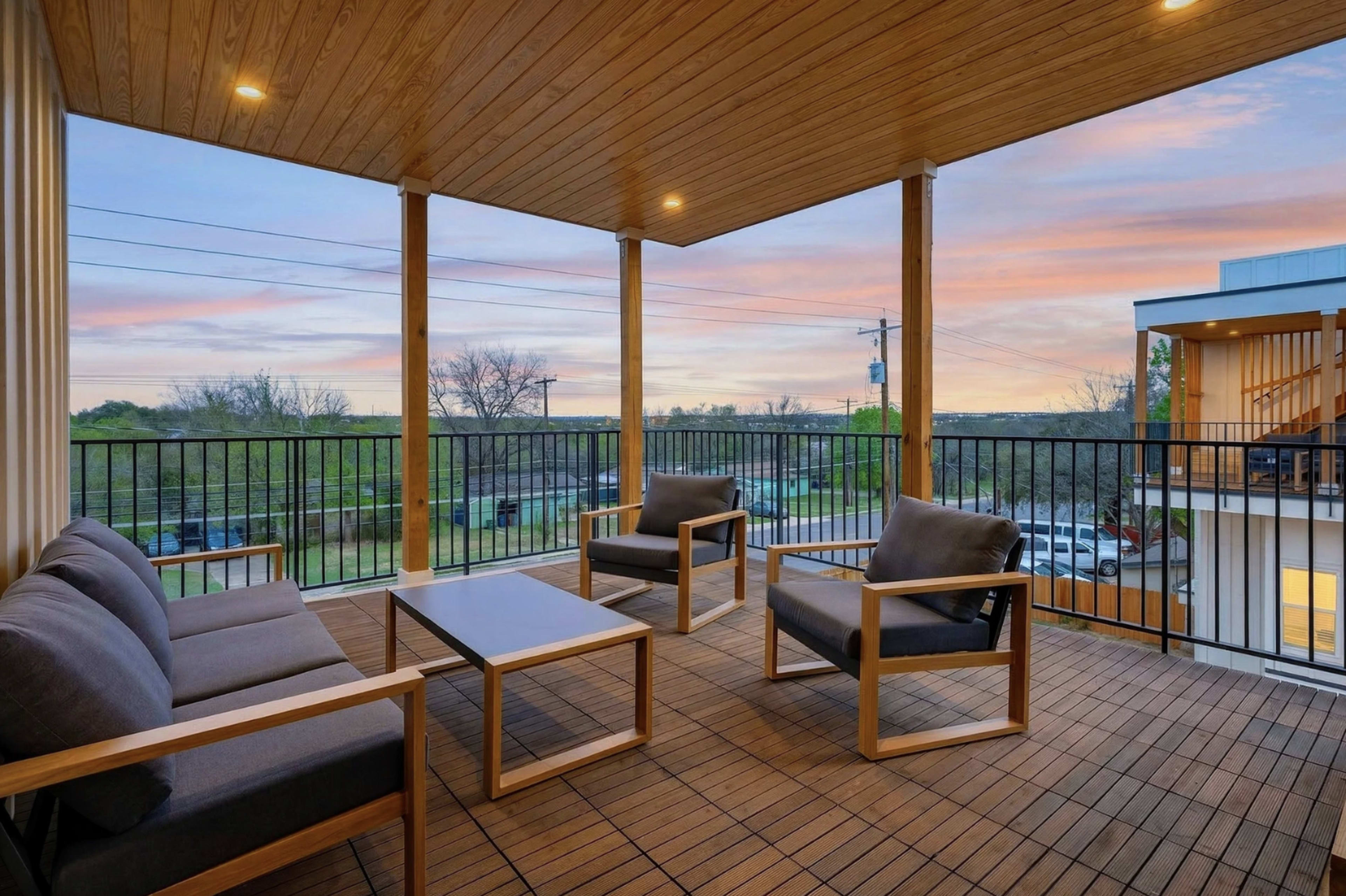 The image shows a spacious outdoor patio with wooden flooring, featuring a seating area with gray cushions and a table, set against a backdrop of a colorful sunset.