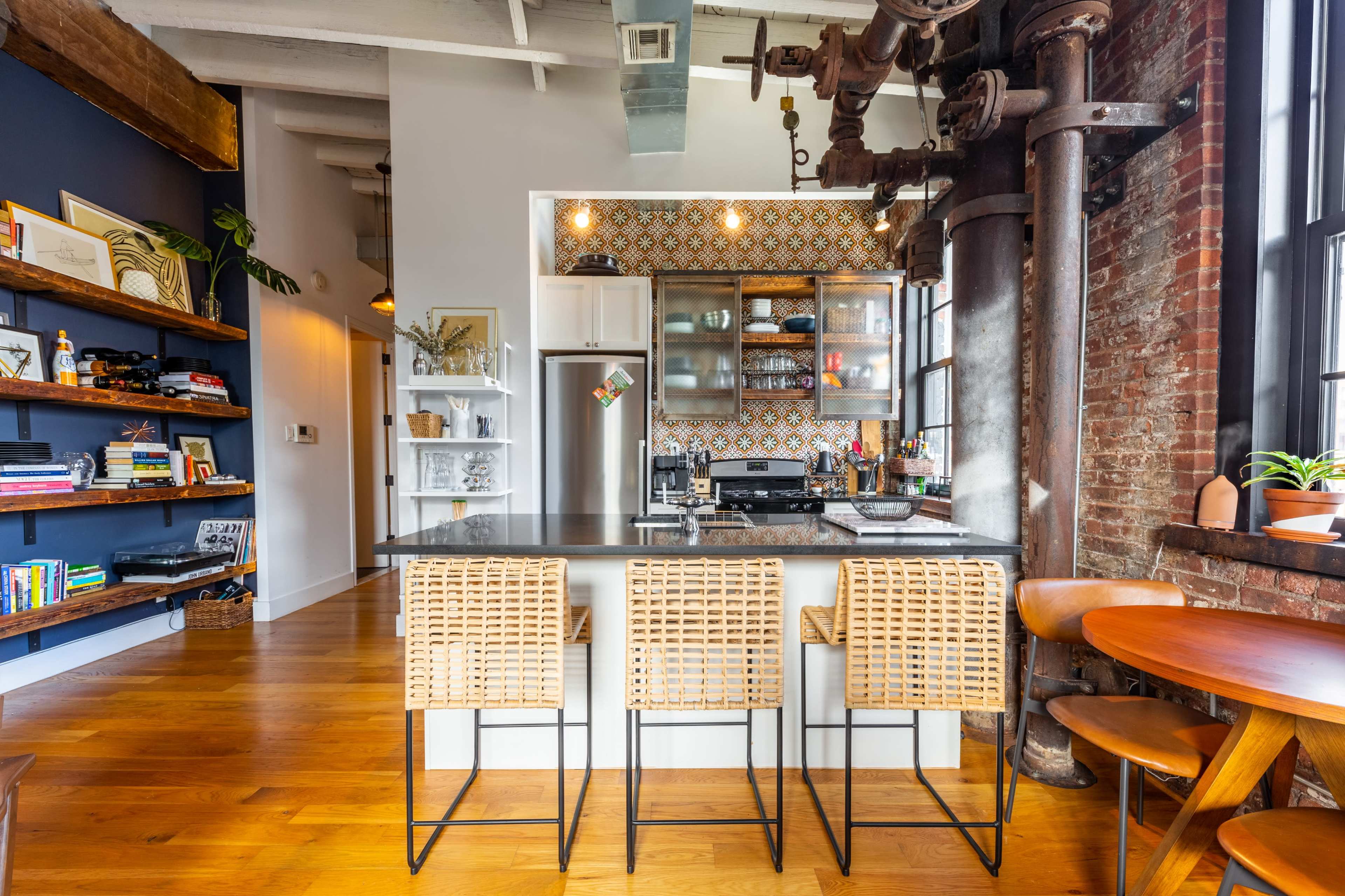 A modern kitchen with a bar island featuring rattan stools, surrounded by exposed brick walls and a patterned backsplash.