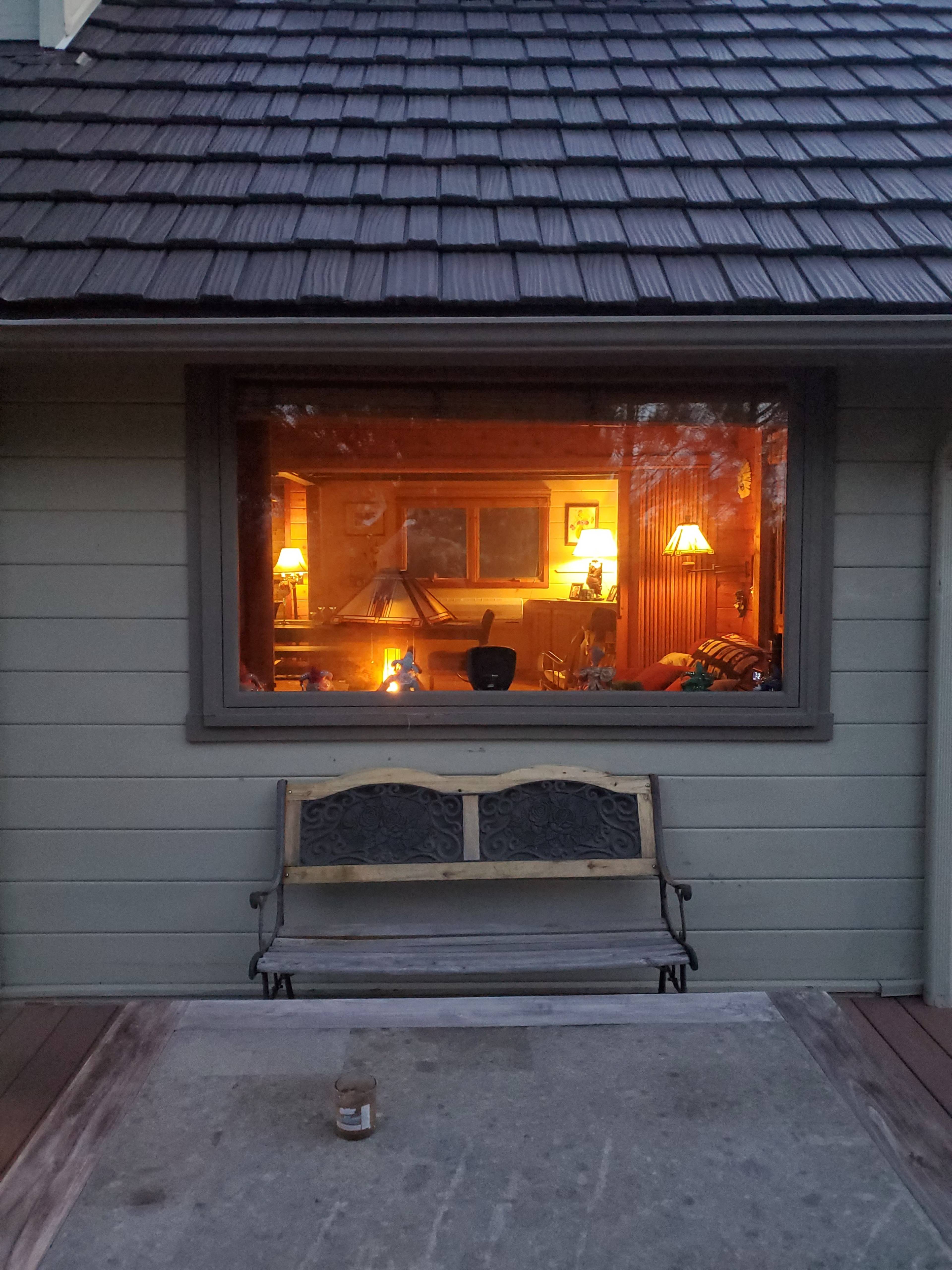 A wooden bench sits on a concrete slab outside a house with a warmly lit interior visible through a large window.