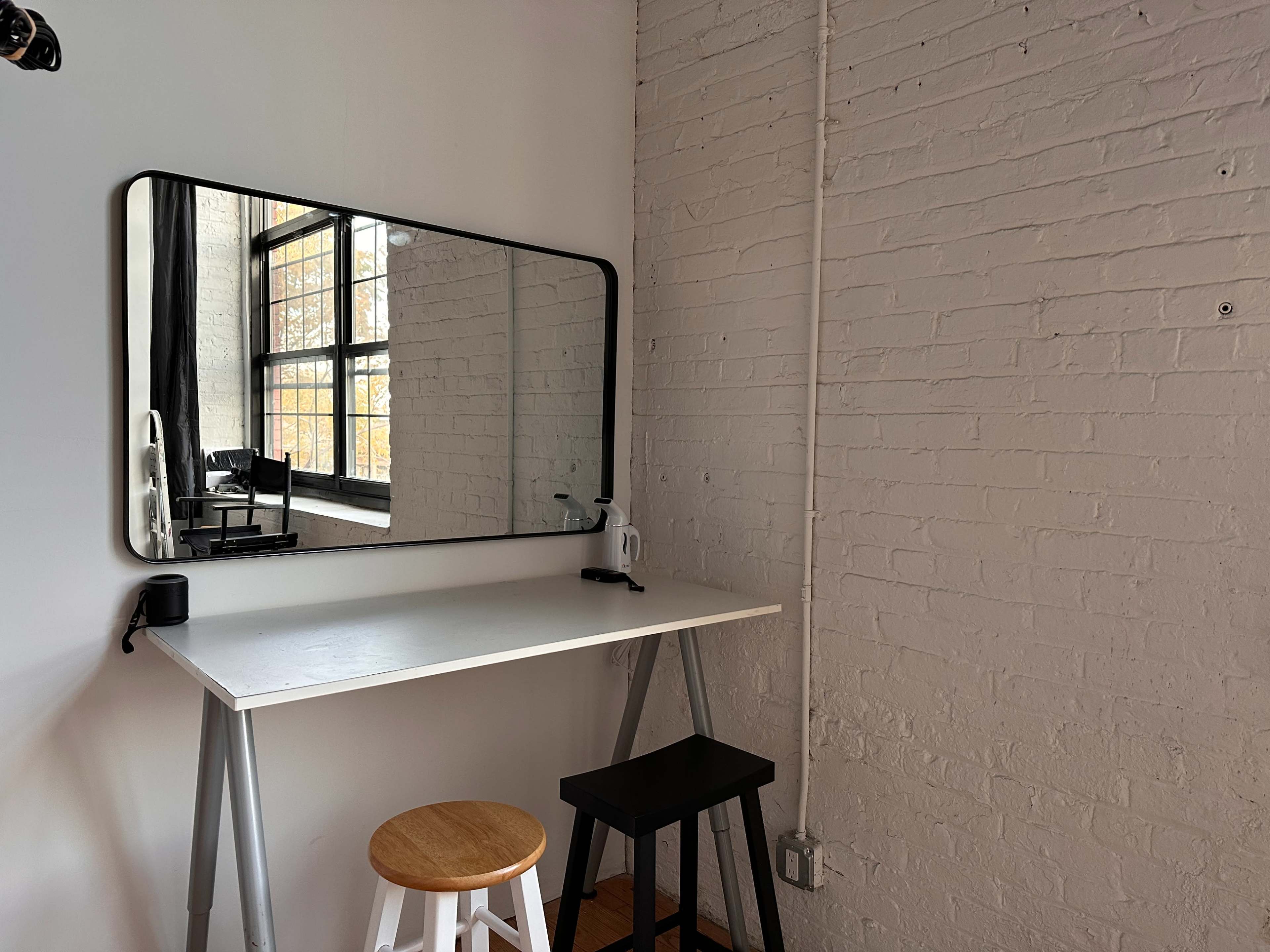 A minimalistic workspace features a large mirror above a white table with two stools, set against a wall of exposed white brick.
