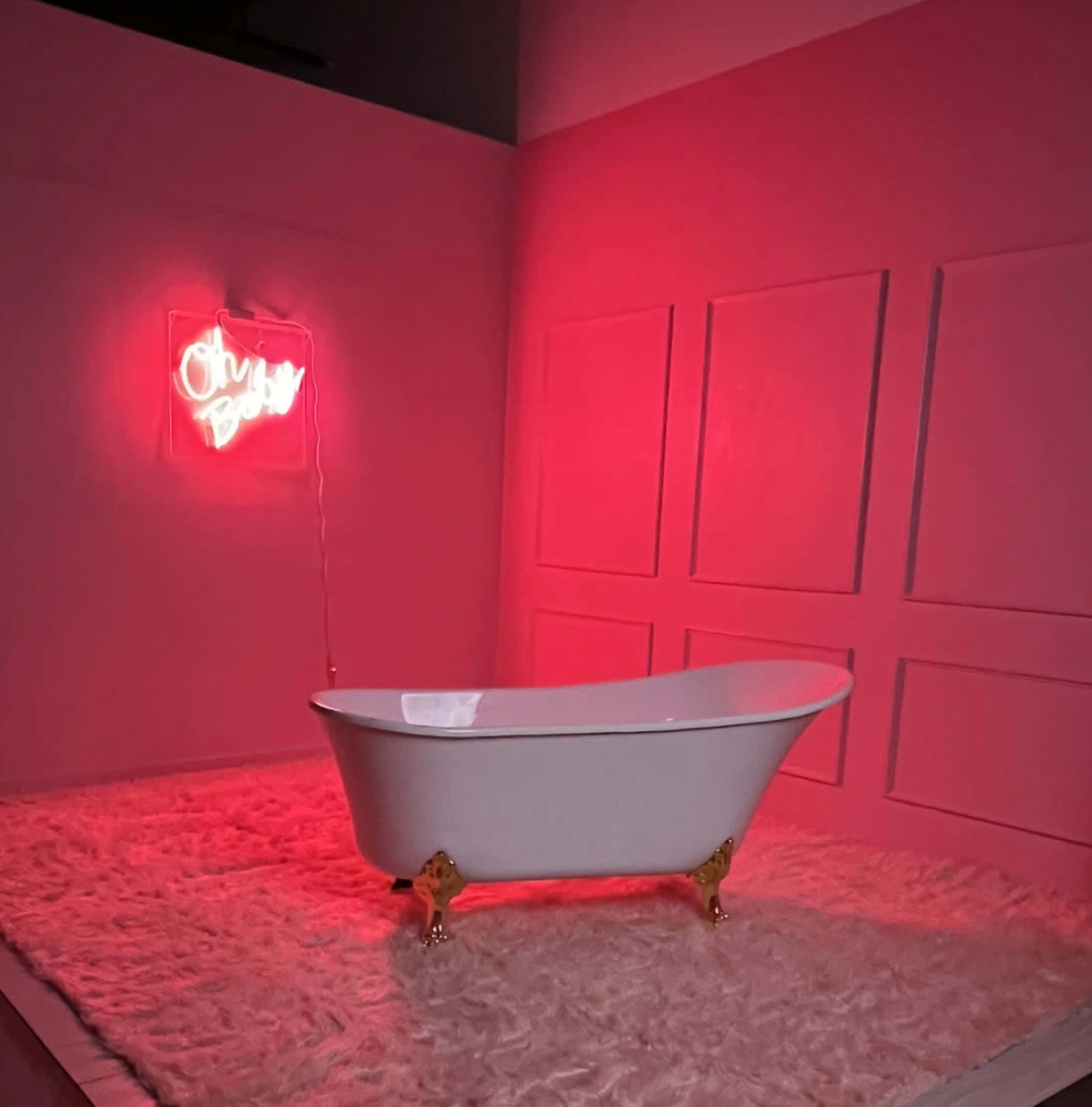 A white freestanding bathtub with golden feet is placed on a plush surface beneath a pink neon sign that says "Oh Baby."