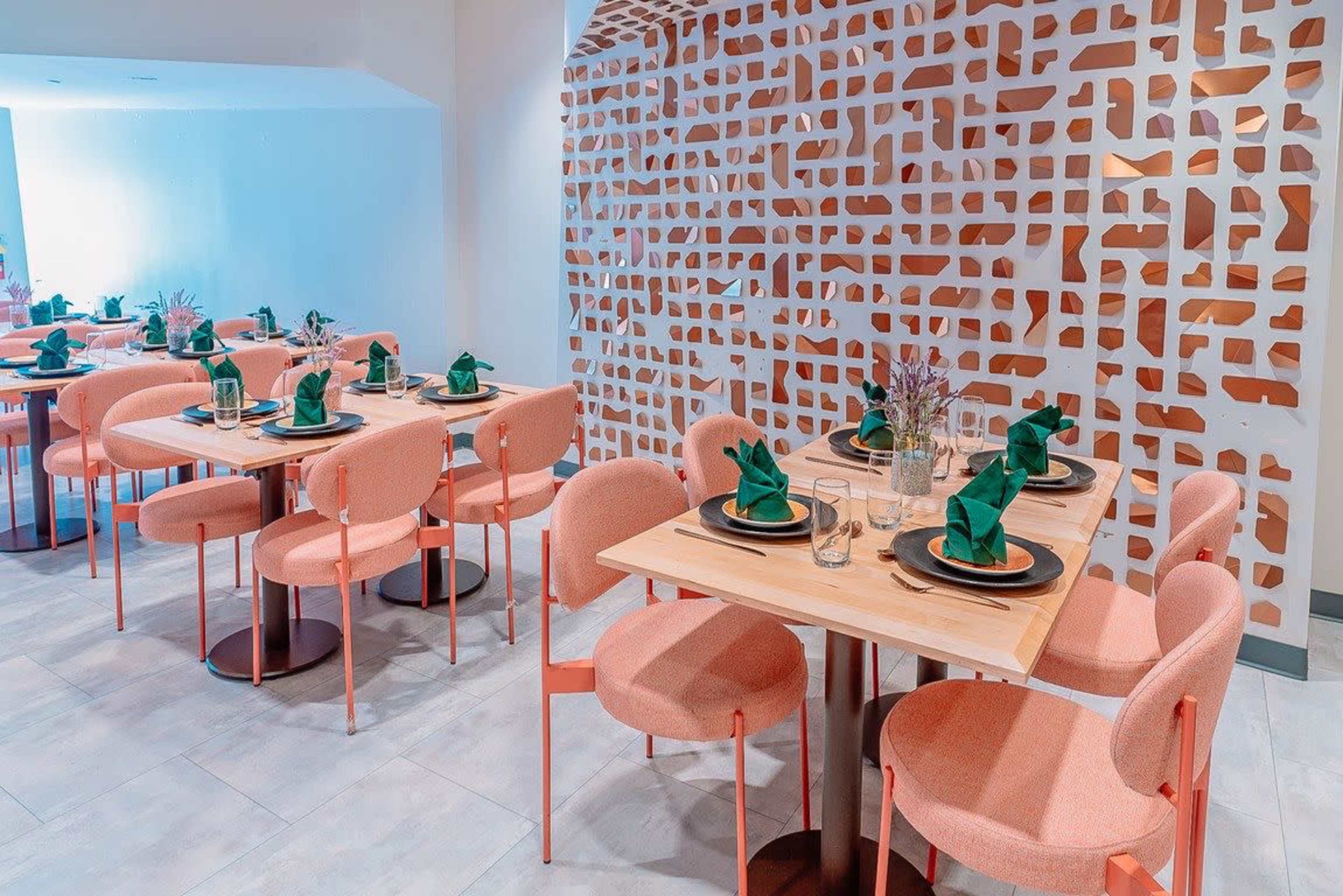 The image shows a modern dining area with several tables set for meals, featuring pink chairs and green napkins against a decorative wall.