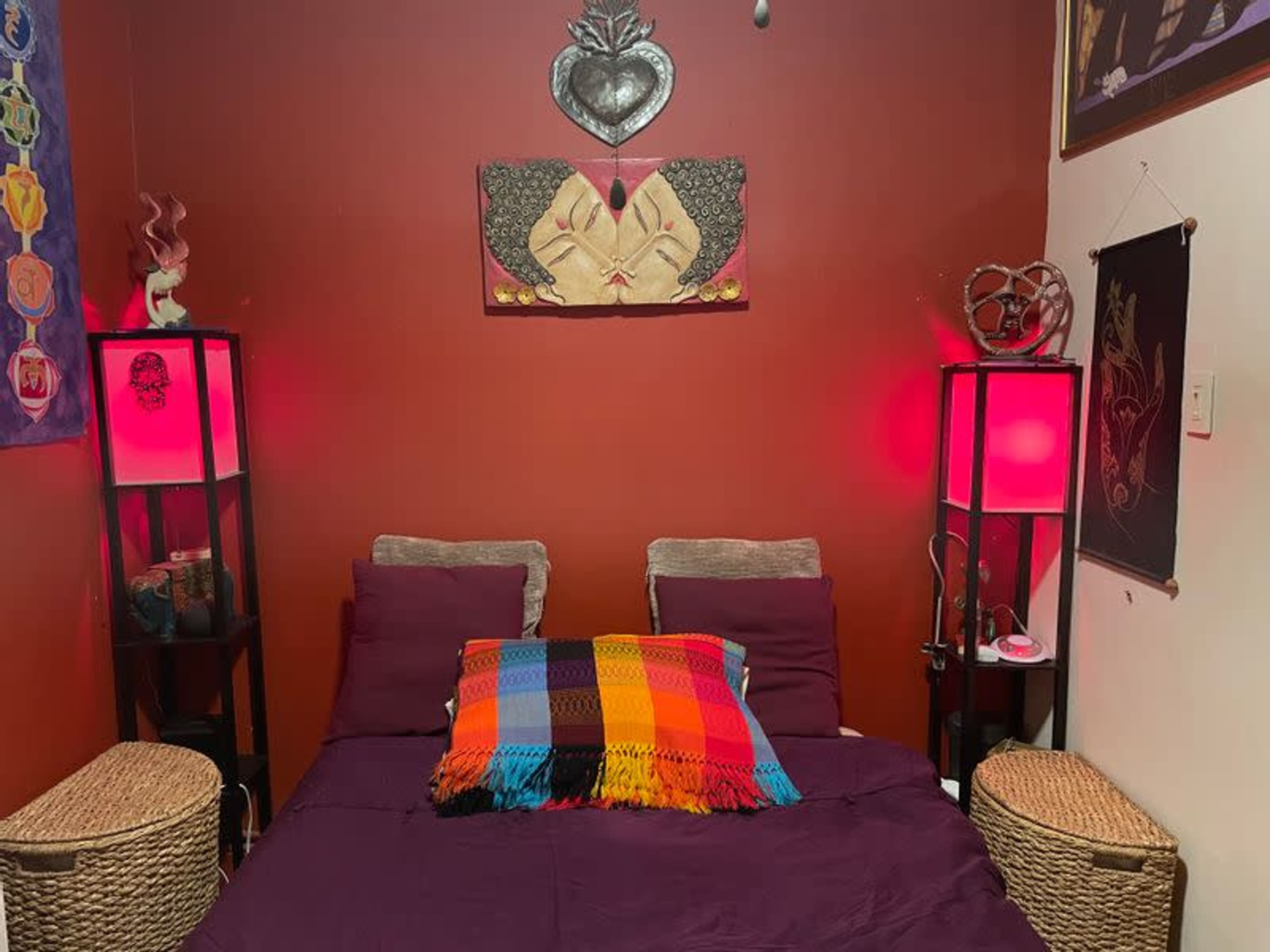 The image shows a small bedroom with a purple bedspread, colorful decorative pillows, and two nightstands with red lamps on either side.